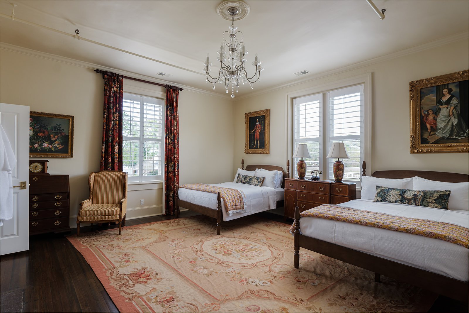 A bedroom with two beds, a chandelier, two windows with shutters, a dresser, a chair, and wall art.
