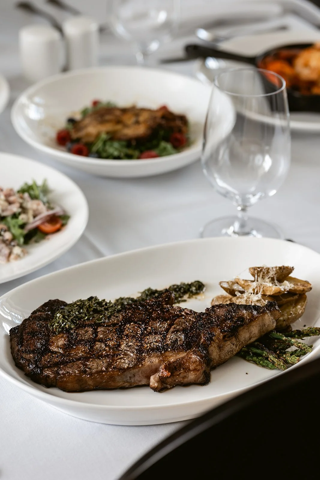 A dinner plate with grilled steak, asparagus, and mushrooms in the foreground, with a glass of water and other dishes in the background.