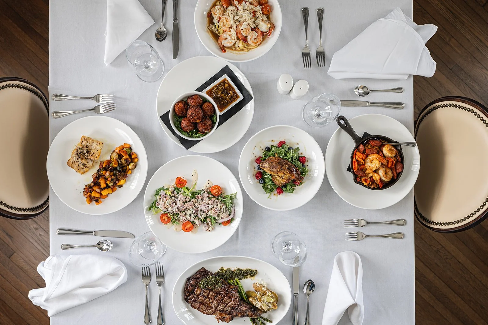A formal dinner table with six plates of food including salads, shrimp, steak, and appetizers, along with silverware, folded white napkins, and empty wine glasses on a white tablecloth.