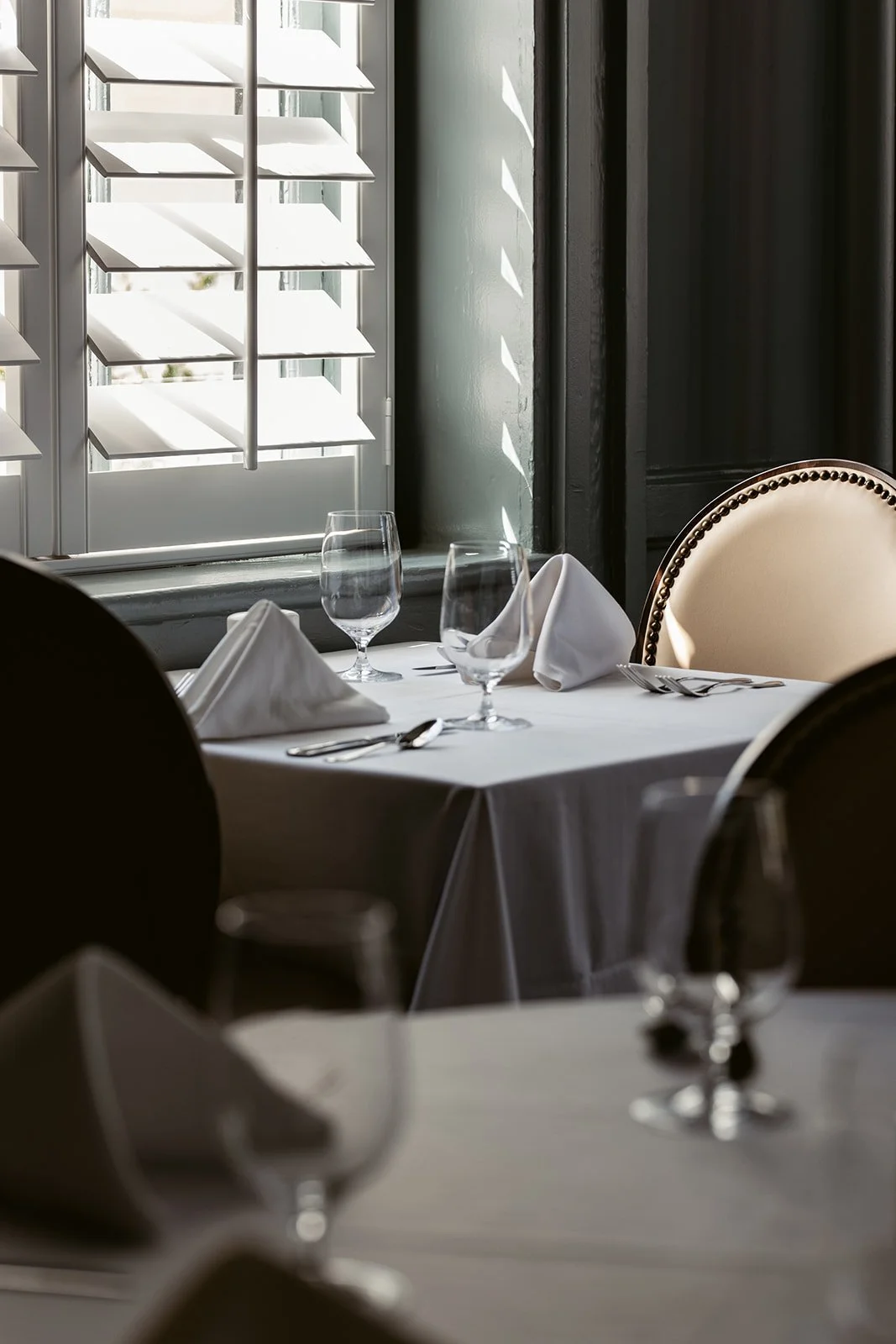 A restaurant table set with white tablecloth, folded white napkins, wine glasses, and silverware near a window with white shutters, sunlight casting shadows on the wall.