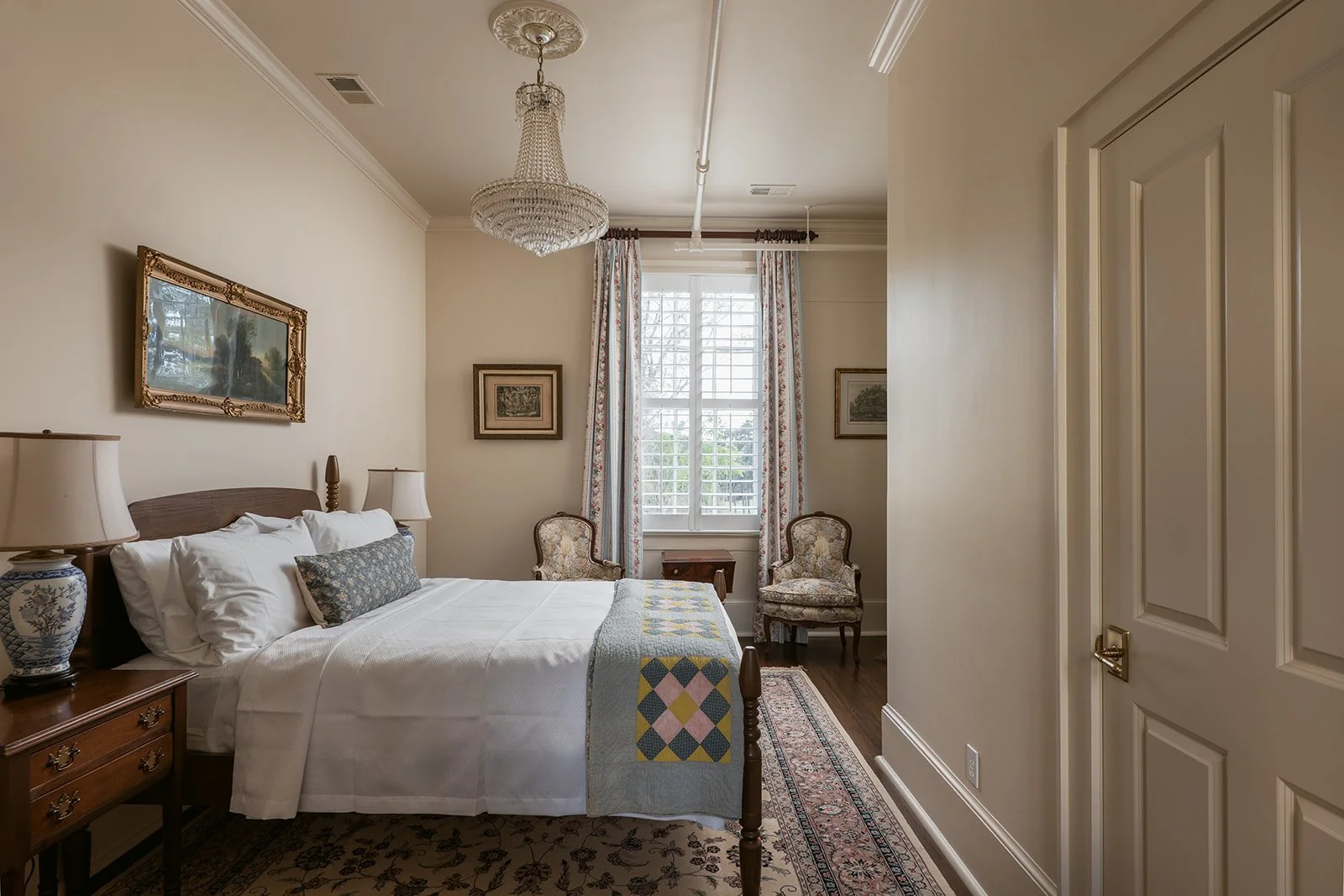 A vintage bedroom with a wooden bed and white bedding, two floral armchairs, a window with floral curtains, a chandelier, and framed pictures on the walls.