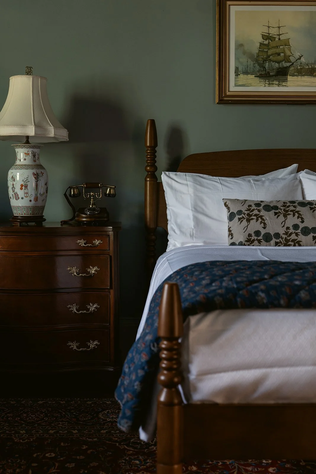 A bedroom corner featuring a wooden bed with white pillows and a patterned throw, a wooden nightstand with a vintage lamp and an old-fashioned rotary phone, and a framed painting of a sailing ship on the wall.