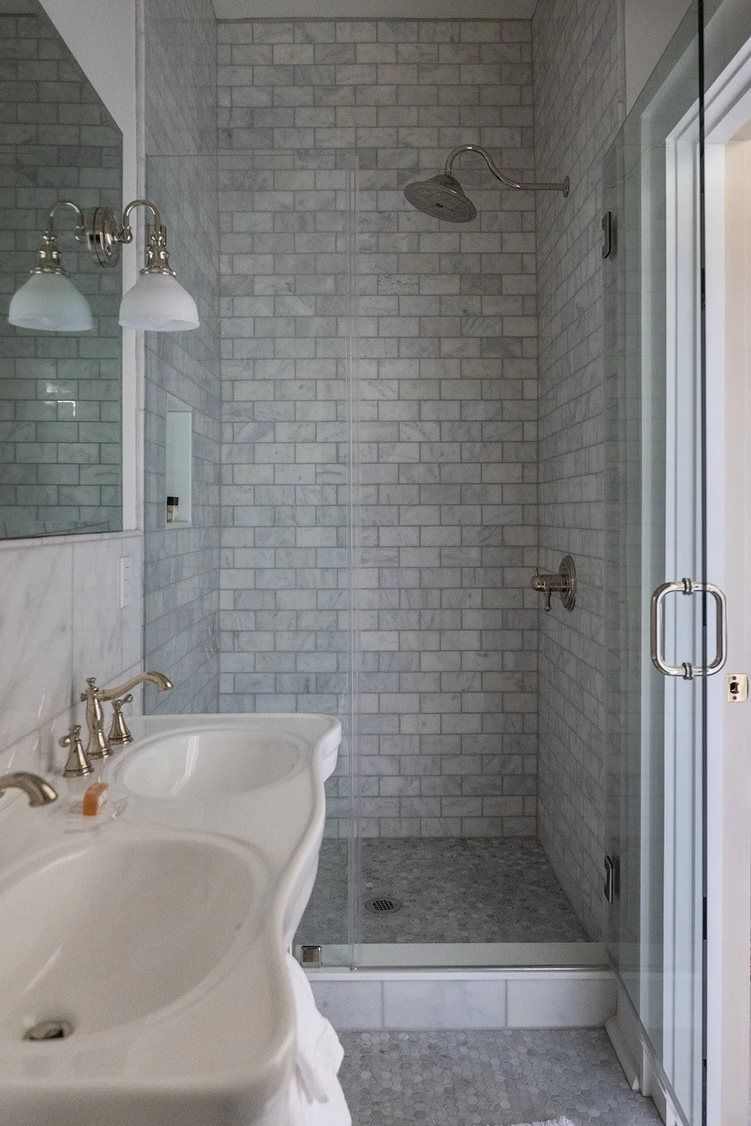Bathroom with a glass-enclosed shower featuring gray brick tiles, a silver showerhead, and a silver faucet with a white double sink, mirror, and wall-mounted light fixtures.