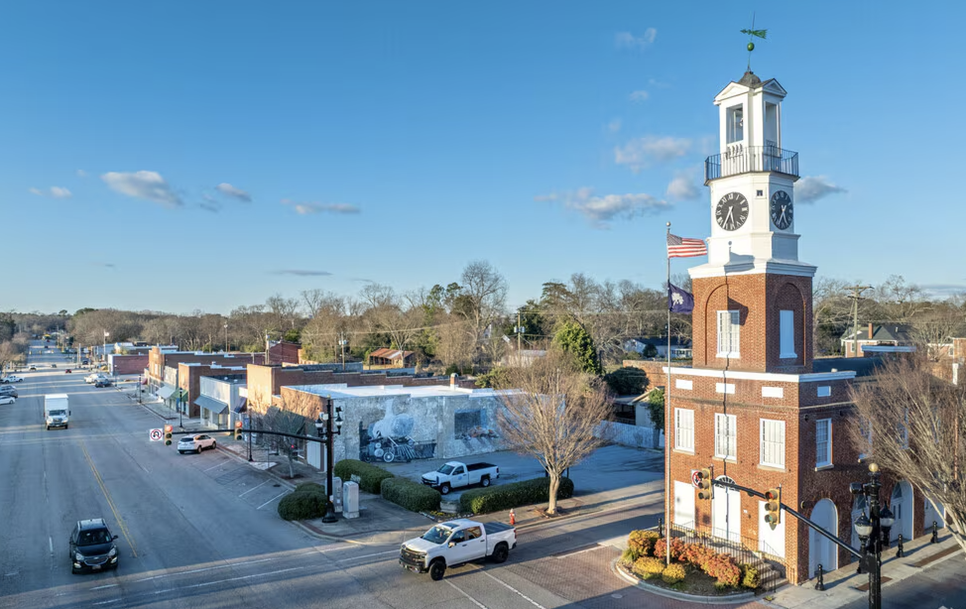 An aerial view of a small town with a prominent white clock tower with a flag and weather vane on top, against a bright blue sky with a few clouds. The town has low-rise buildings, trees, and a few cars on the streets.