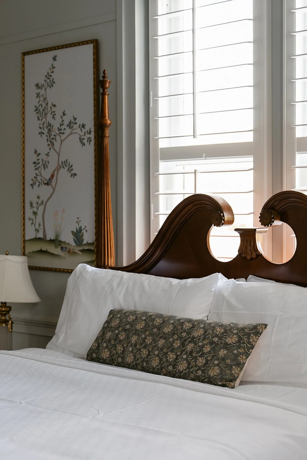 Close-up of a bed with wooden headboard, white pillows, decorative pillow, and a lamp beside with window shutters in the background. Hotel Everett | Boutique Hotel in Winnsboro, SC, historic hotel