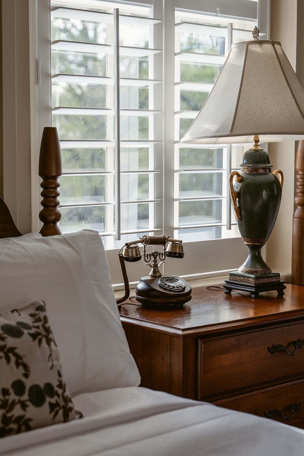 A cozy bedroom corner with a wooden nightstand holding a vintage rotary telephone and a large decorative lamp with a beige shade, situated in front of a window with white plantation shutters.