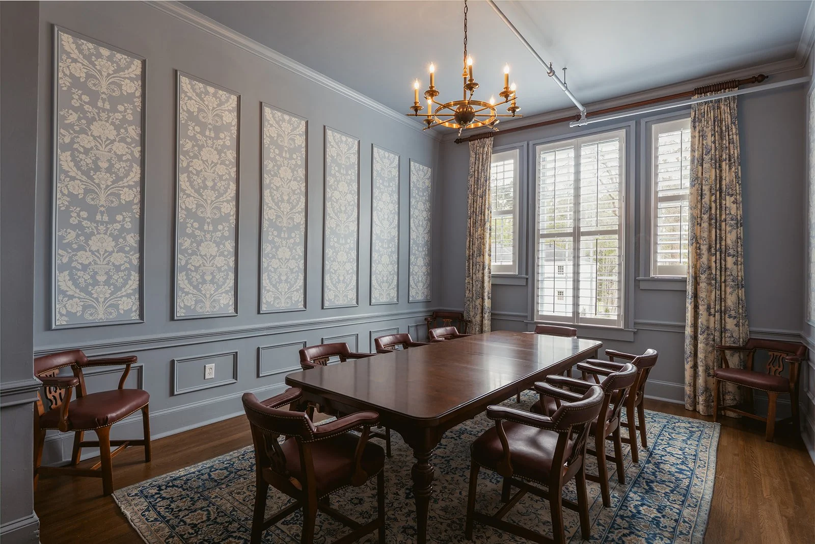 A formal dining room with a large wooden table and ten leather chairs. The room features light blue walls with decorative moldings, a brass chandelier, three large windows with plantation shutters, patterned drapes, and a patterned area rug on hardwood floor.