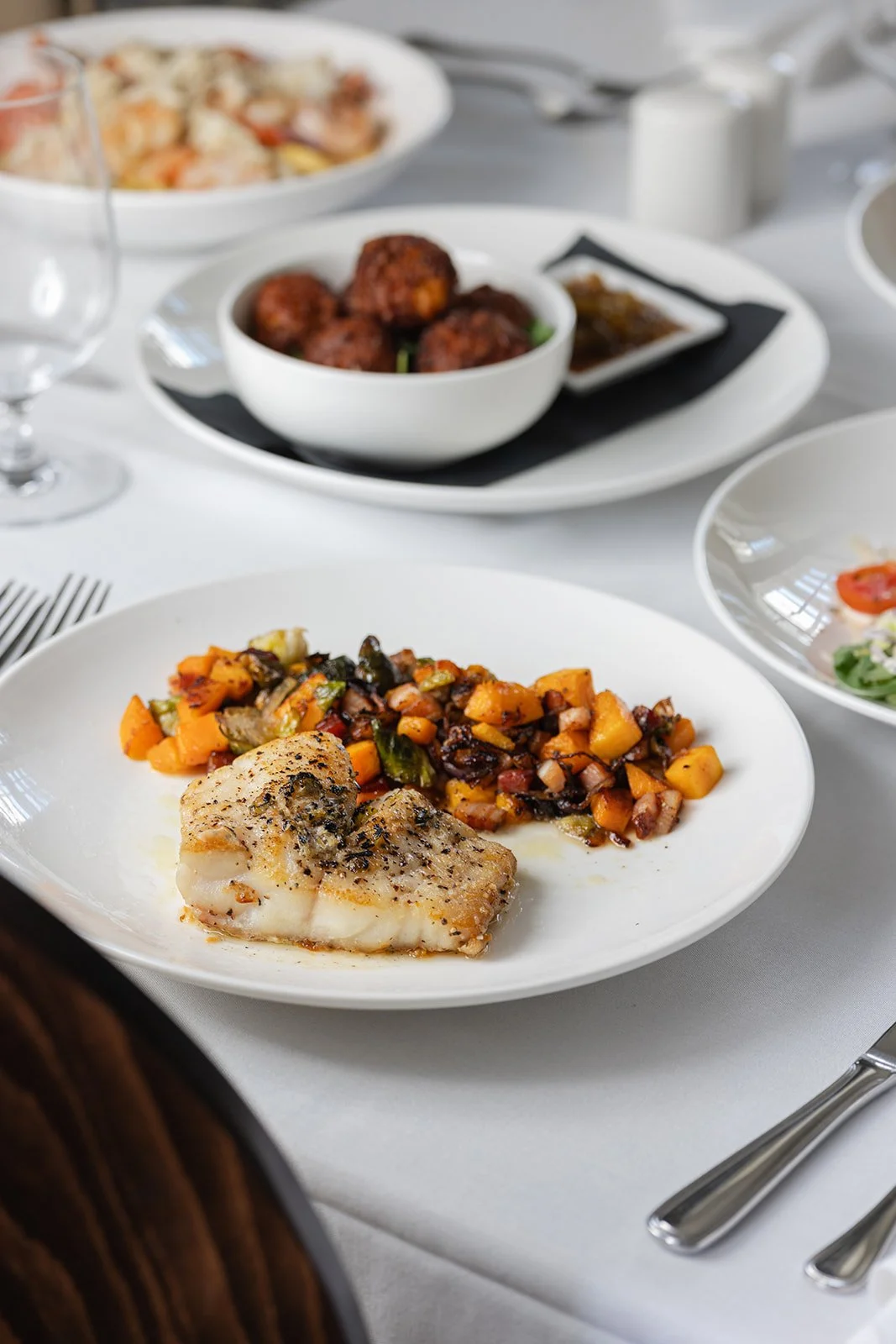 A white plate with a piece of seasoned fish fillet and a serving of roasted vegetable medley on a white tablecloth.