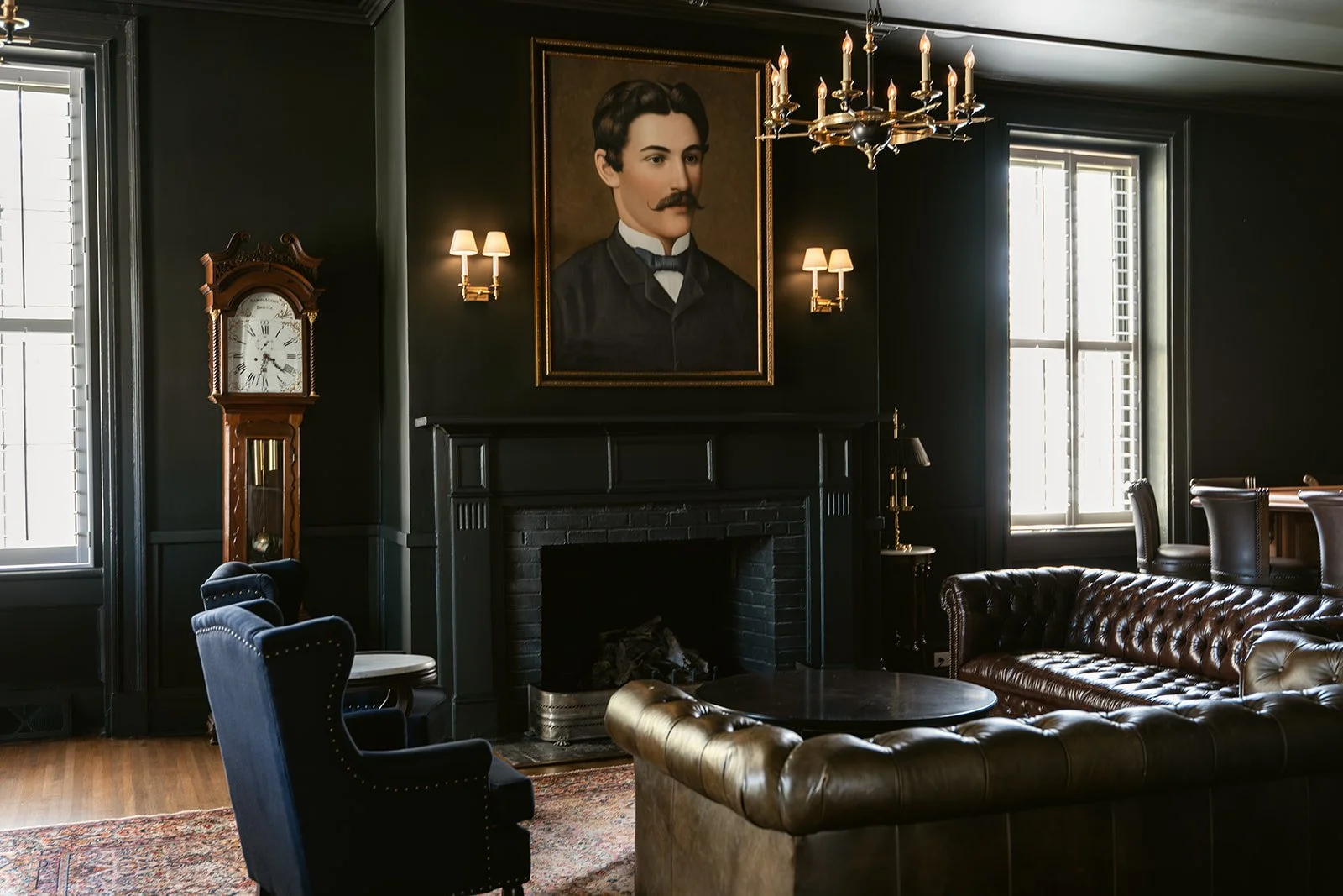 A richly decorated lounge with black walls, featuring a portrait of a man with a mustache, two windows with white shutters, a wooden grandfather clock, a dark leather chesterfield sofa, a round black coffee table, a tall lamp, and a chandelier.