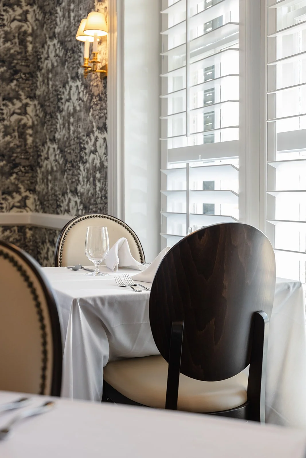 Elegant restaurant table setting with white tablecloth, wine glass, napkin, and silverware, near white window shutters and a wall with black and white patterned wallpaper, illuminated by a wall-mounted lamp.