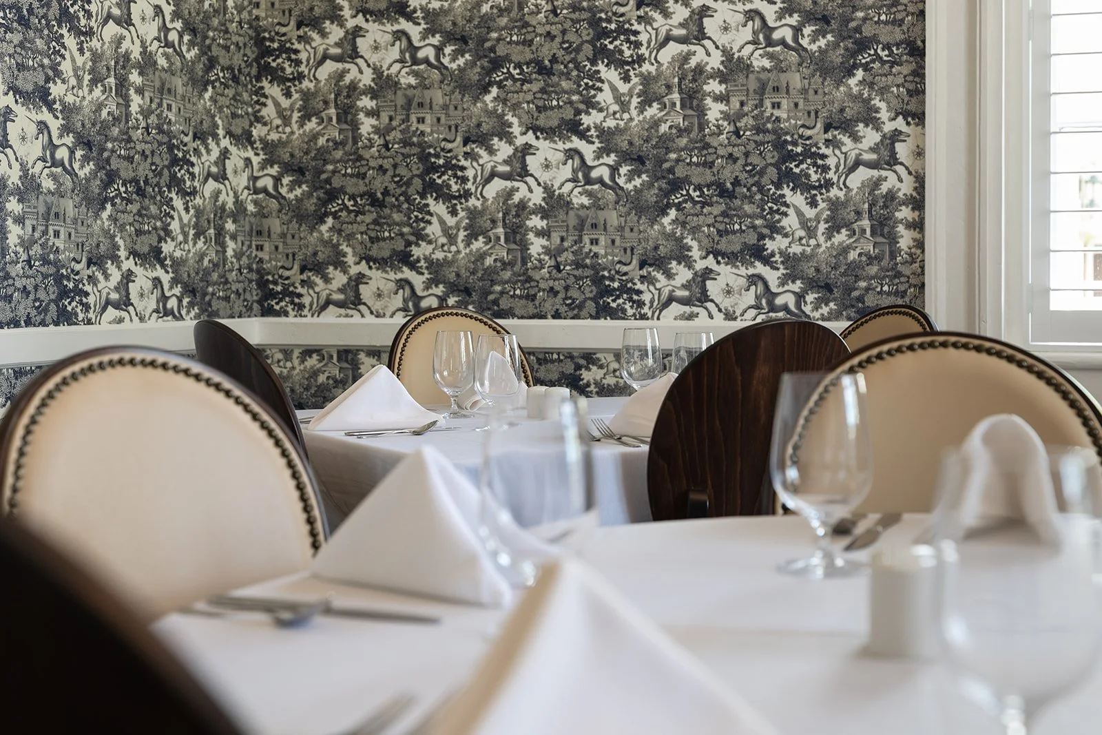A formal dining room with a round table set with white napkins, glasses, and silverware, featuring elegant upholstered chairs and a black-and-white toile wallpaper with images of horses, trees, and houses.