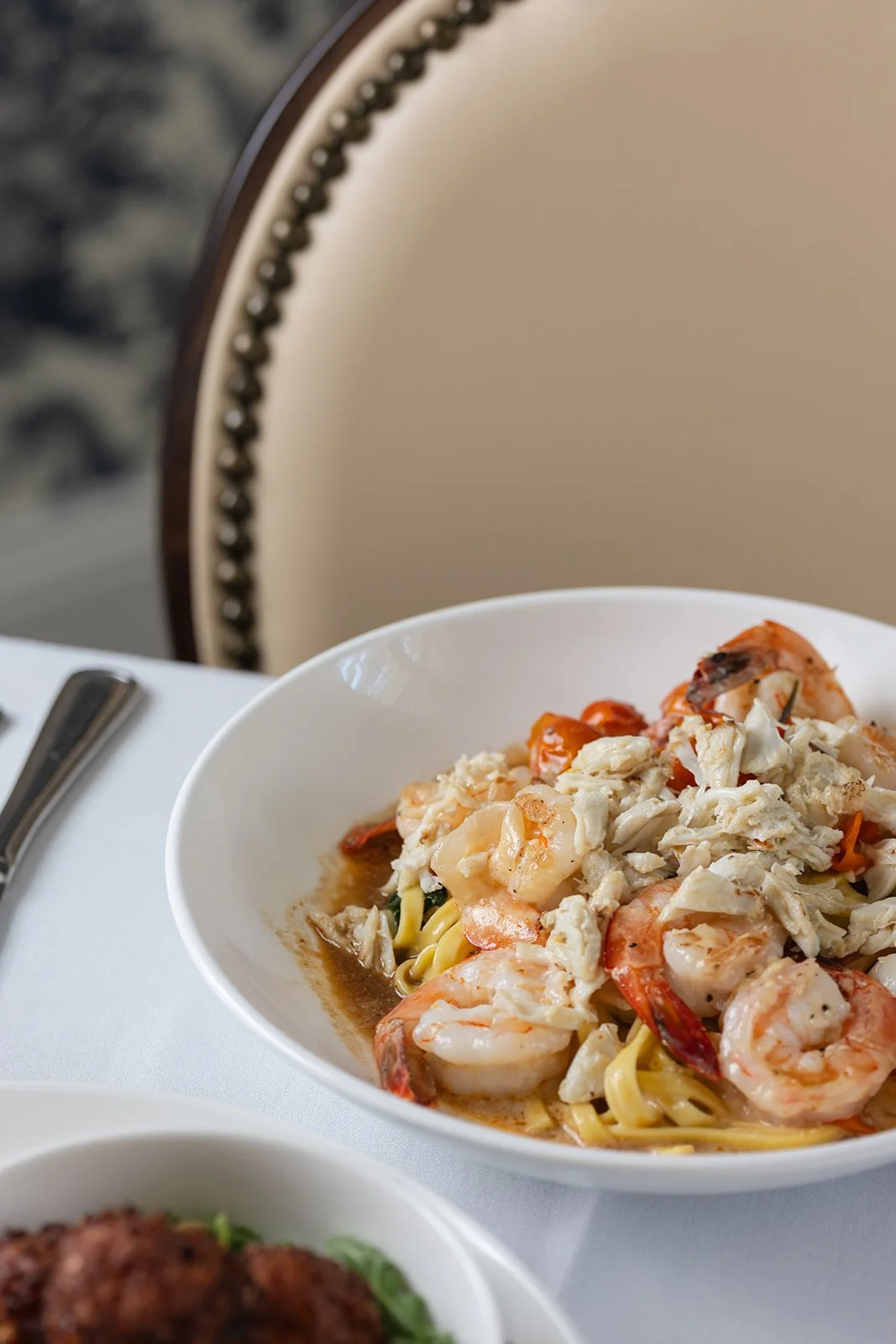 A bowl of seafood pasta with shrimp, crab, and cherry tomatoes on a white table, with a blurred background showing a beige chair with nailhead trim.