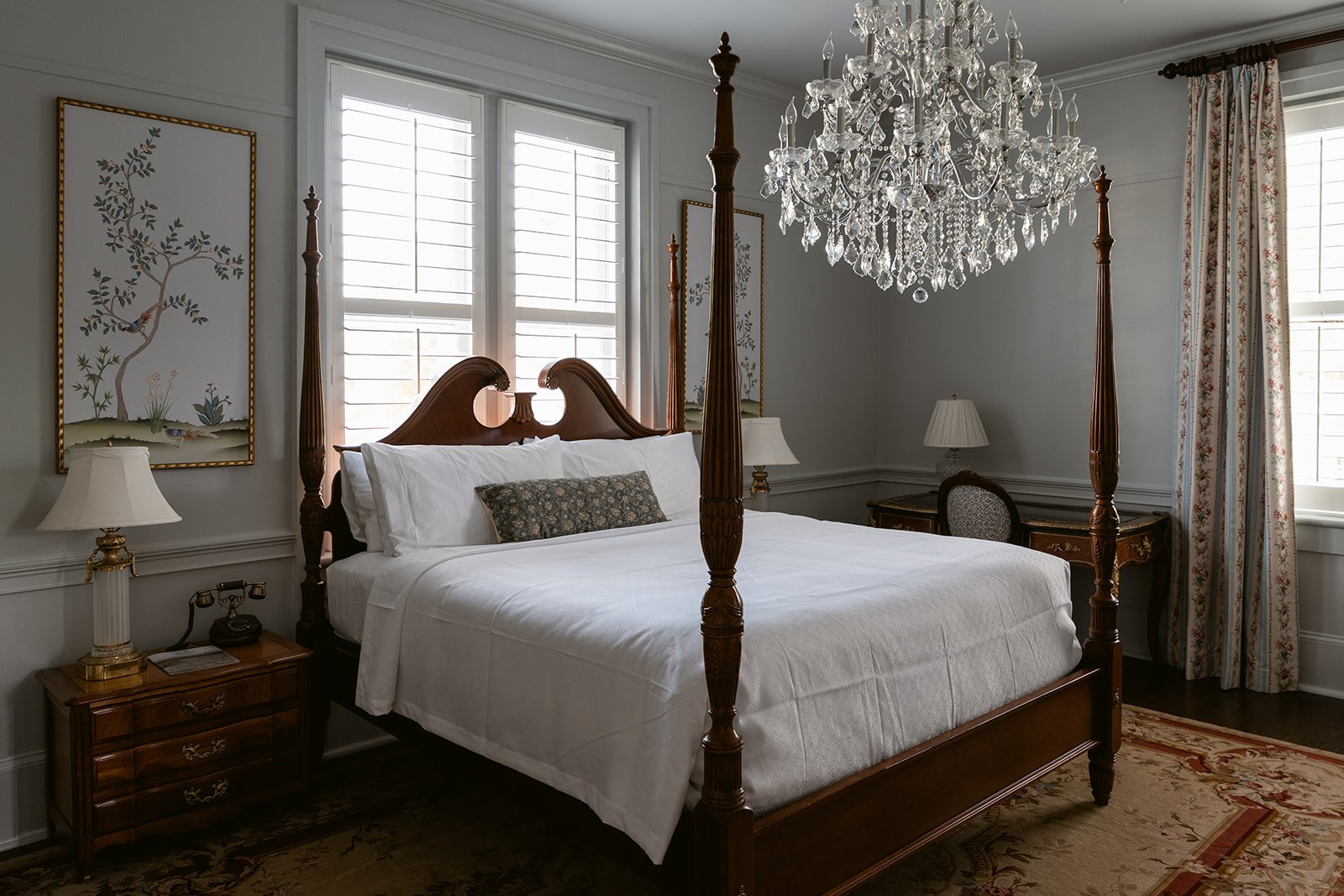A luxurious bedroom with a four-poster bed, white bedding, and pillows, illuminated by natural light from large windows. The room features elegant chandeliers, classic wooden furniture, framed artwork on the walls, and floral curtains.