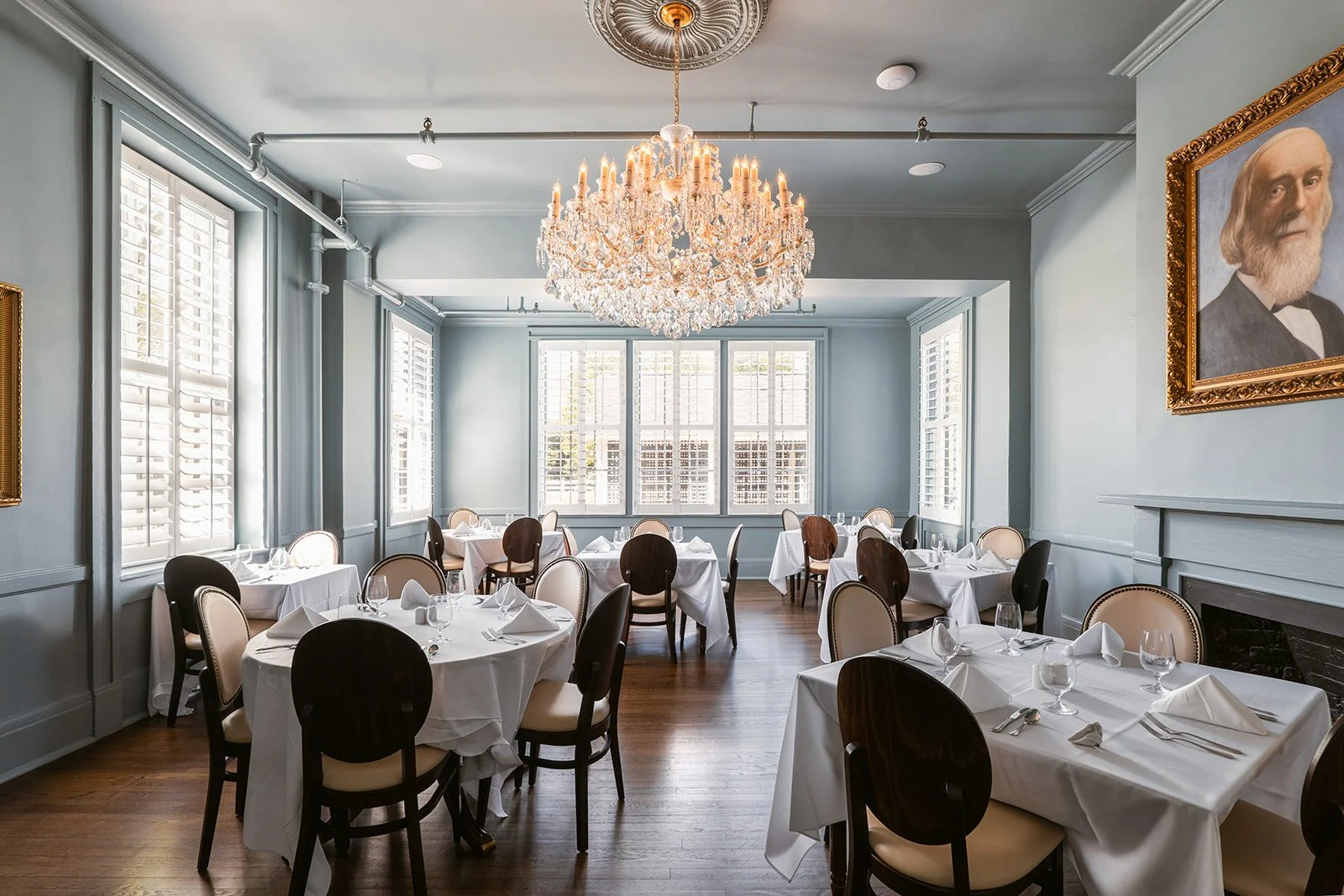 Elegant restaurant dining room with white tablecloths, dark and light chairs, a large chandelier, portrait painting, and large windows with shutters.