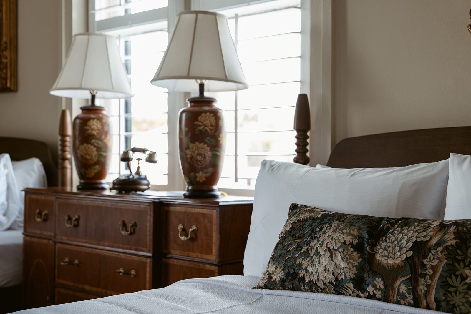 A bedroom with a bed, white pillows, and a decorative pillow with floral patterns. A wooden nightstand with two ornate lamps and a vintage rotary phone. A window with white shutters letting in natural light.