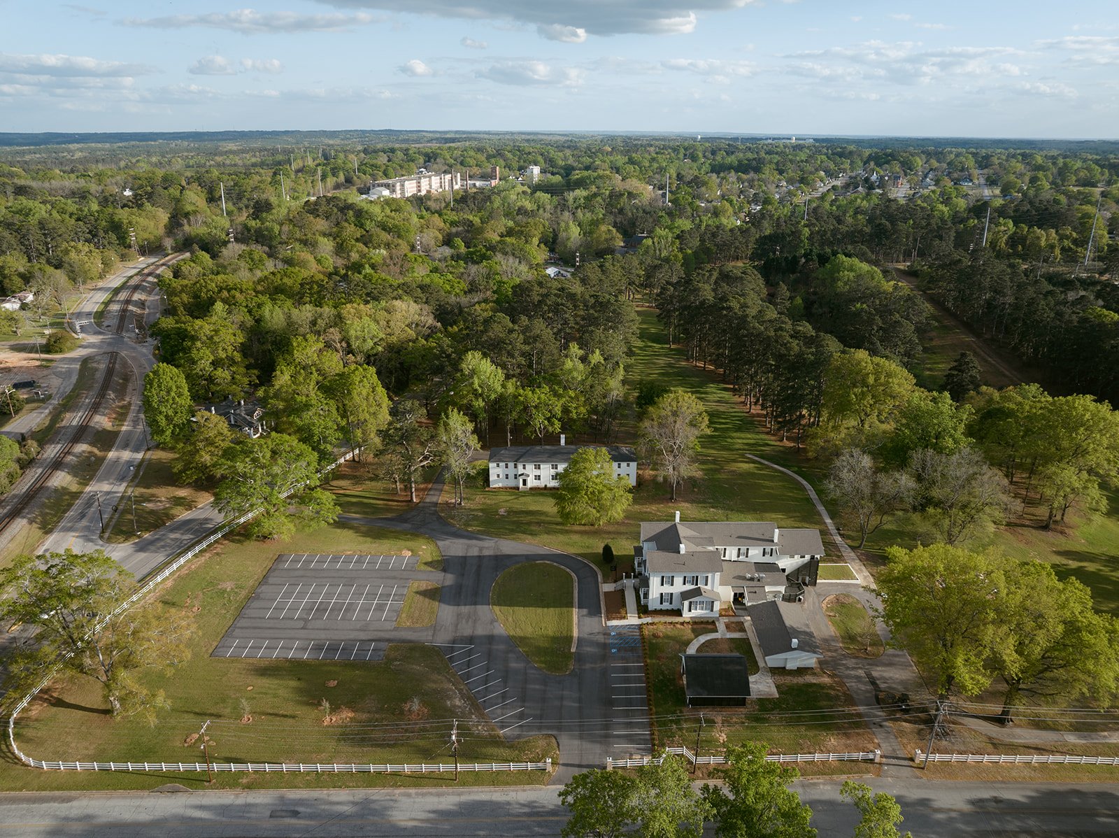 Aerial view of suburban area with houses, trees, parking lots, a railway track, and roads on a partly cloudy day.