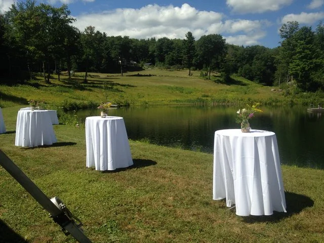 Three round tables with white tablecloths and small flower arrangements on a grassy area near a pond with trees and a cloudy sky in the background.