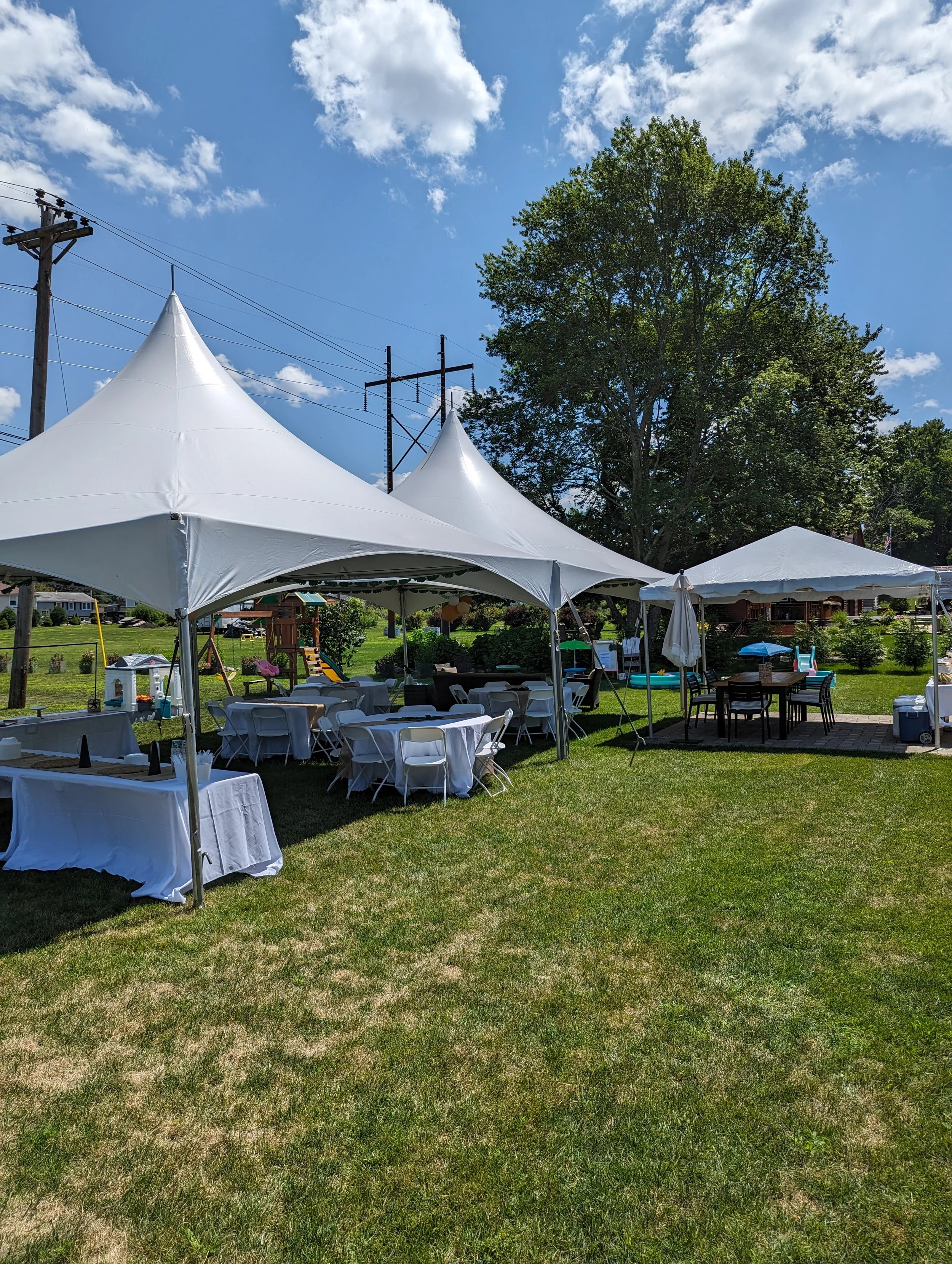 Outdoor event setup with white tents, tables with white tablecloths, chairs, and a playground on a grassy lawn under a blue sky with clouds.