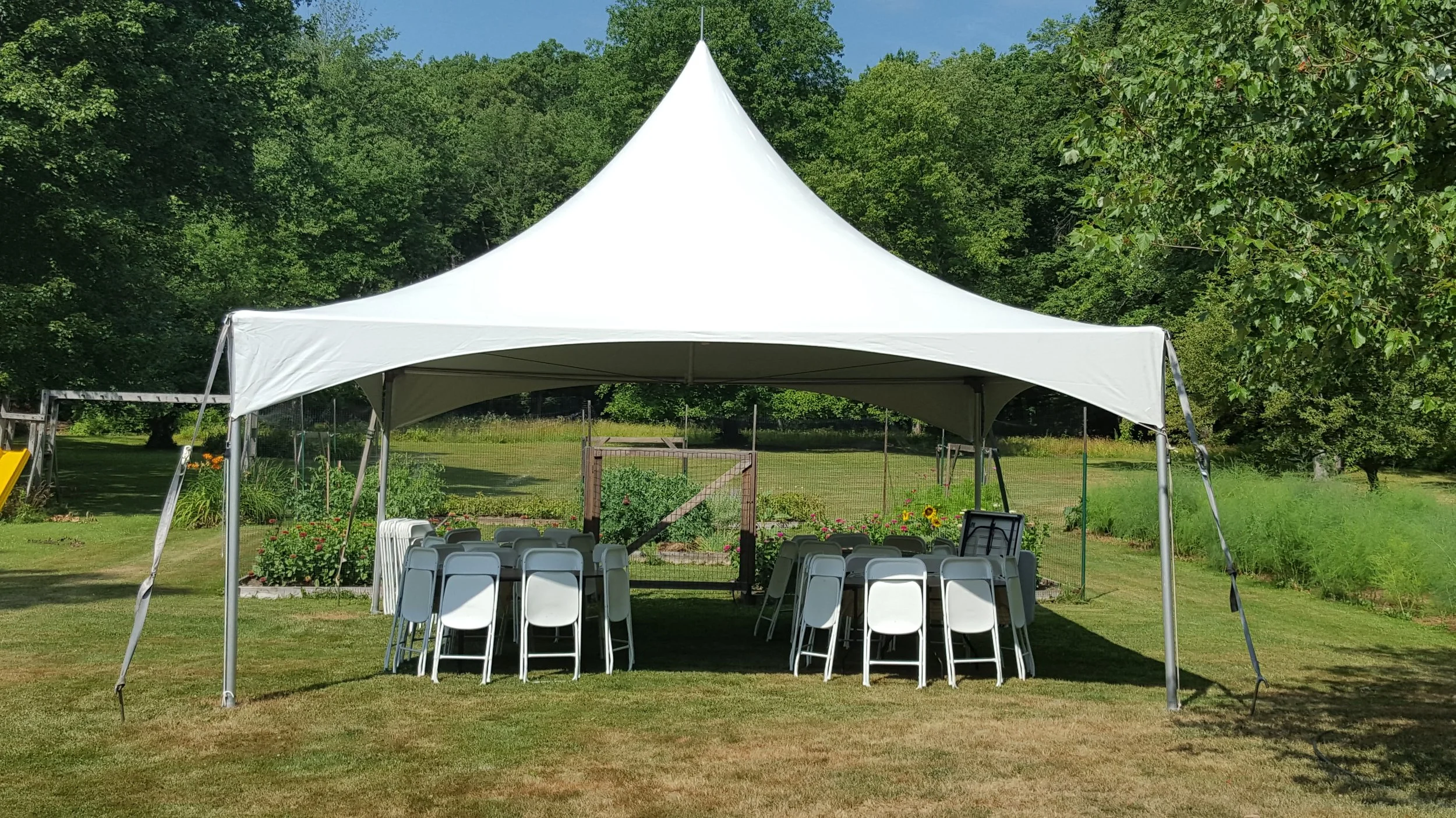White event tent set up outdoors on a grassy area with chairs and tables underneath, surrounded by trees and garden beds.