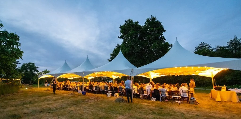 Outdoor evening event under a large white tent with illuminated interior, filled with seated guests and a few standing people, surrounded by trees.