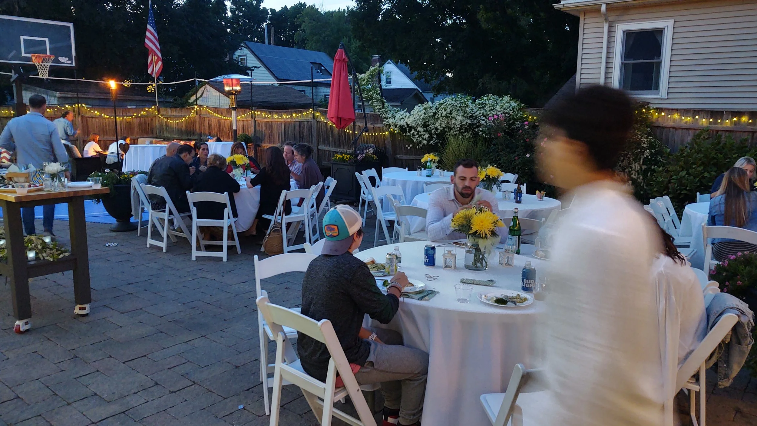 An outdoor evening gathering with people sitting around round tables with white tablecloths, decorated with yellow flowers, and string lights along the fence, in a backyard with a basketball hoop, American flags, and a house.