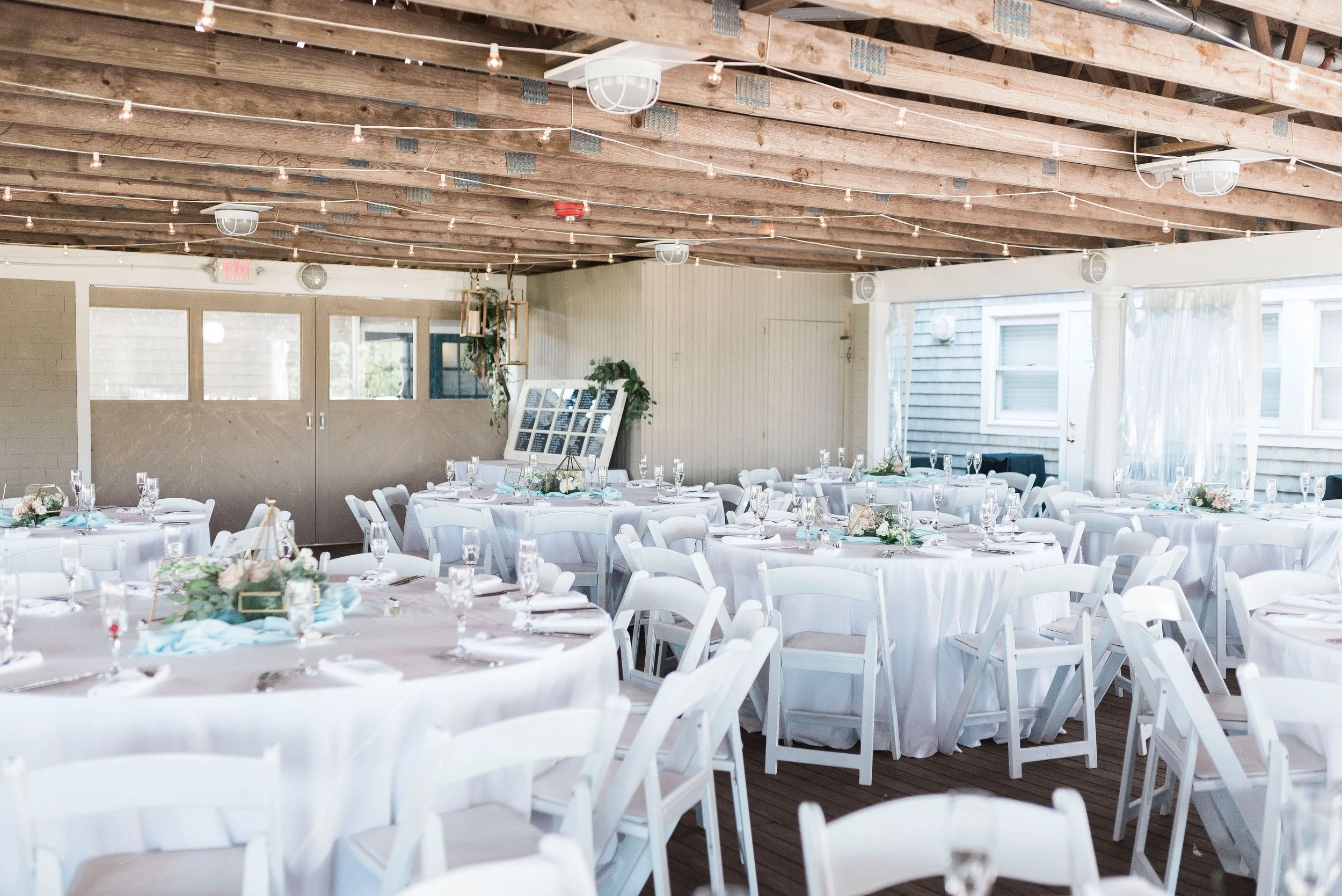 Decorated banquet hall with round tables covered in white tablecloths, set with glassware and napkins, featuring floral centerpieces and light blue accents, illuminated by string lights and natural light from windows.