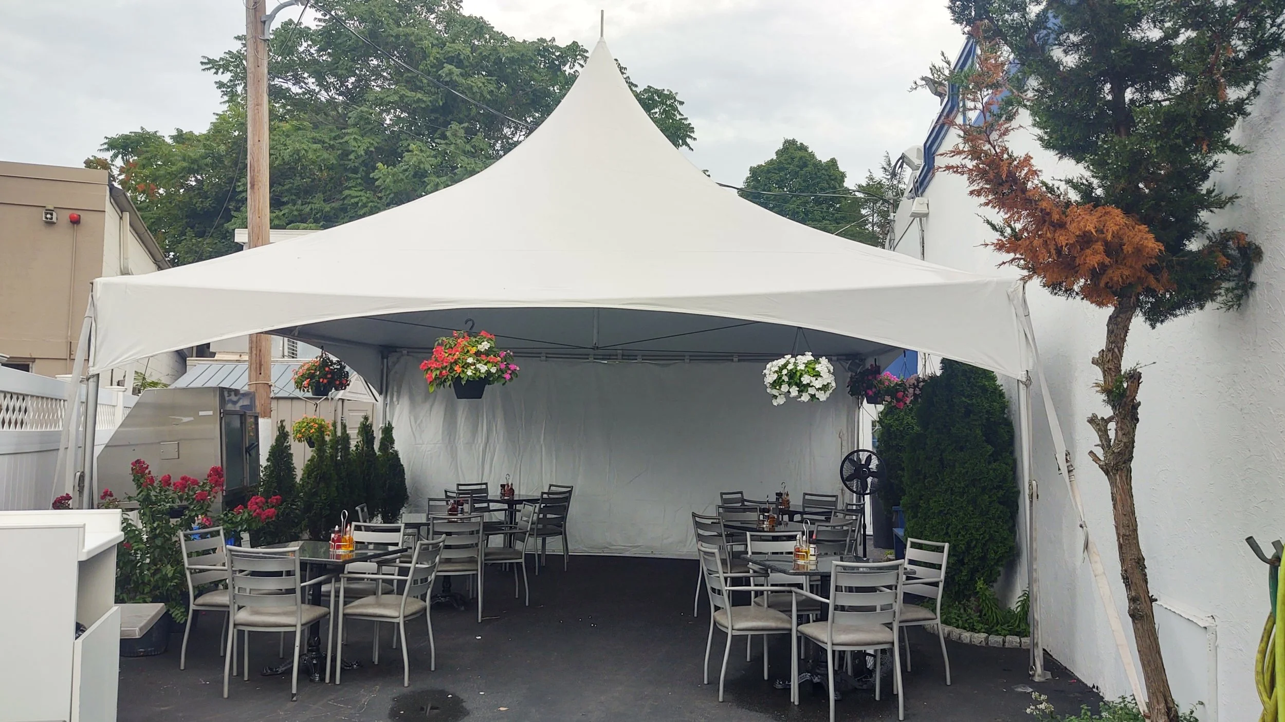 Empty outdoor dining area with tables and chairs under a white tent, decorated with hanging flower baskets, surrounded by potted plants and trees.