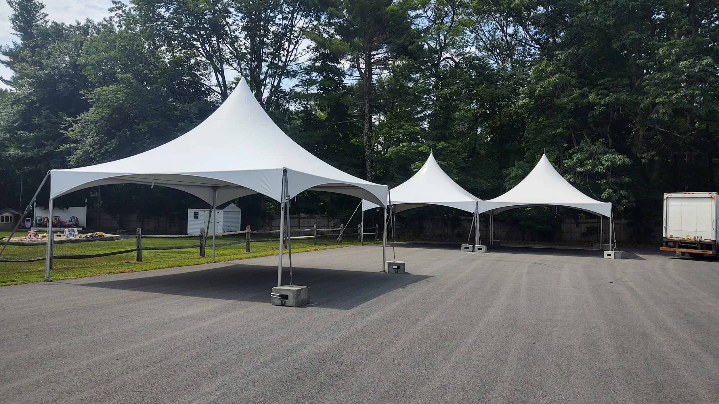 Three white event tents set up outdoors on a gravel surface with a background of trees and a fence.