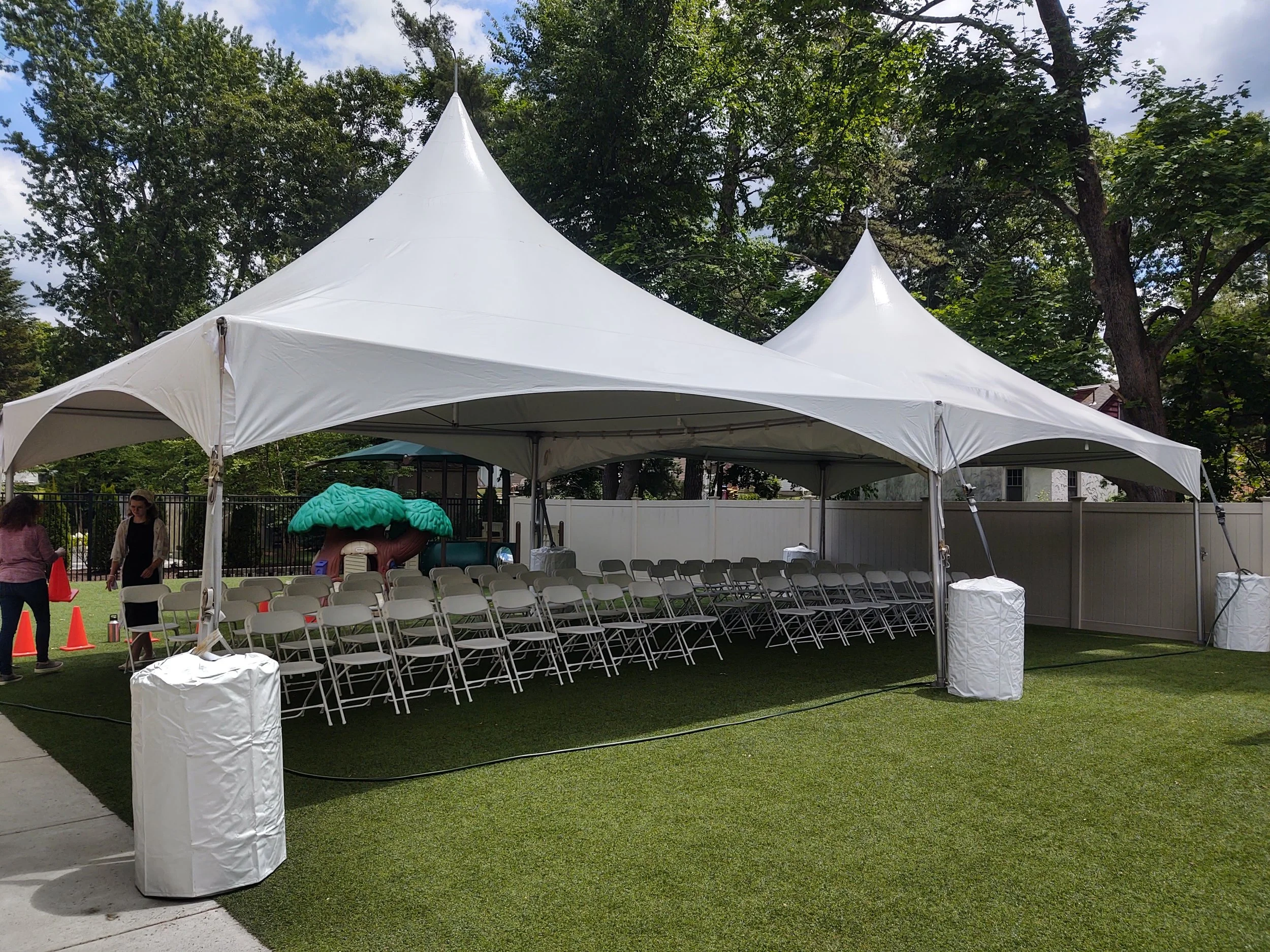 A large white event tent set up outdoors on a grassy area with rolling chairs arranged underneath. There are orange cones and two women on the left side and some trees in the background.