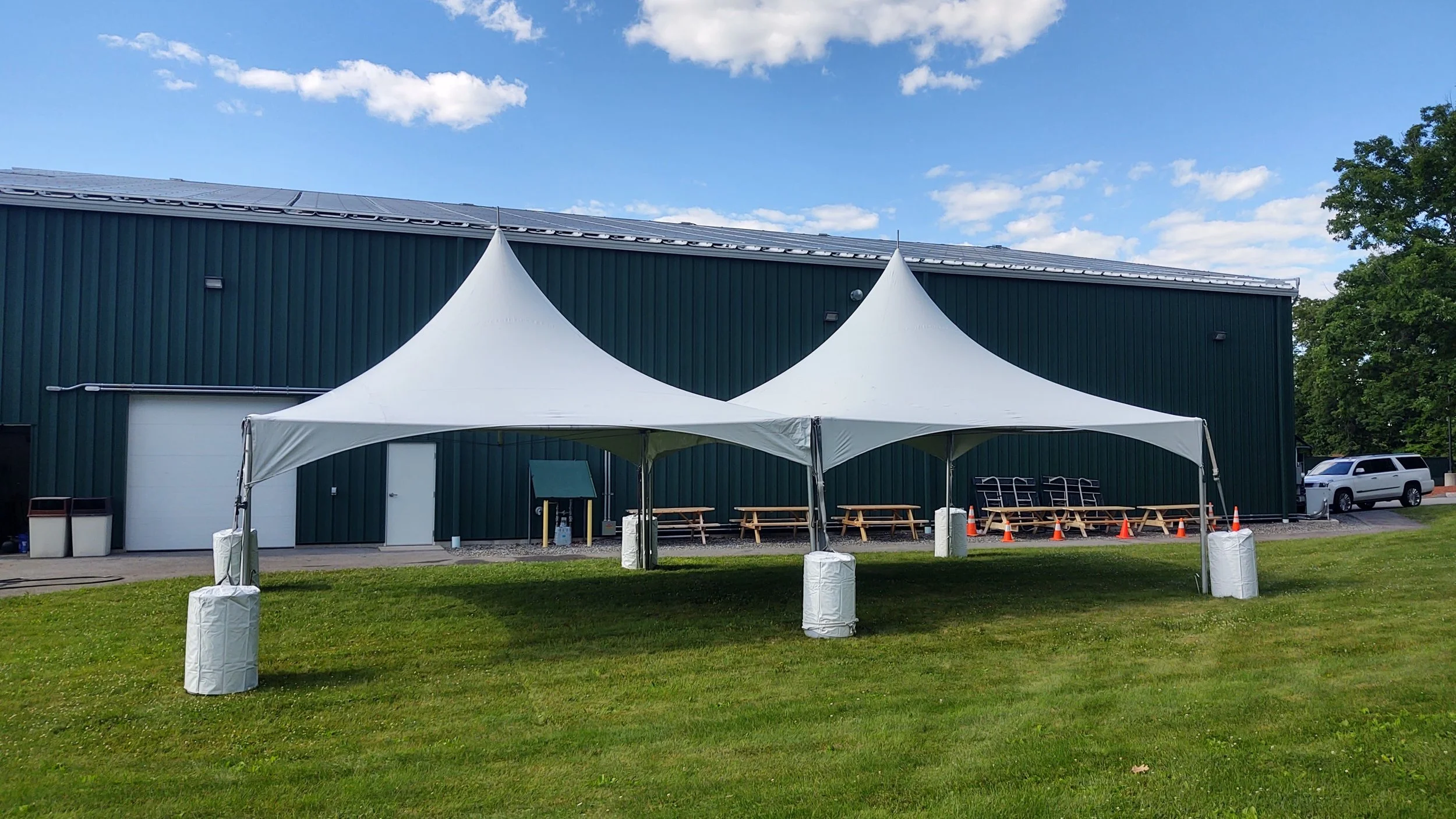 Two white event tents set up on a grassy lawn in front of a green industrial building, with picnic tables under the tents and a vehicle parked on the side.