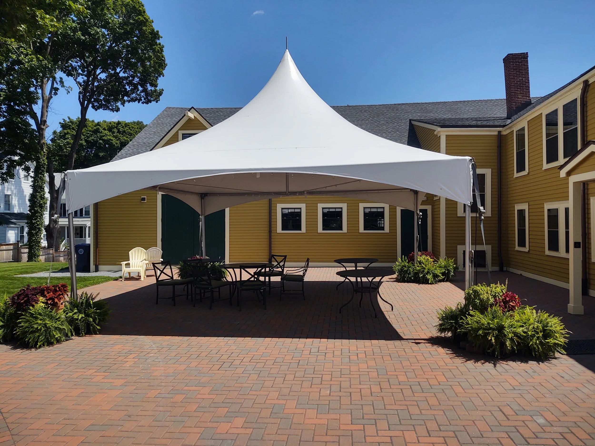 Outdoor patio area with a large white event tent, black metal tables and chairs, surrounded by potted plants, in front of a yellow house with multiple windows, under a clear blue sky.