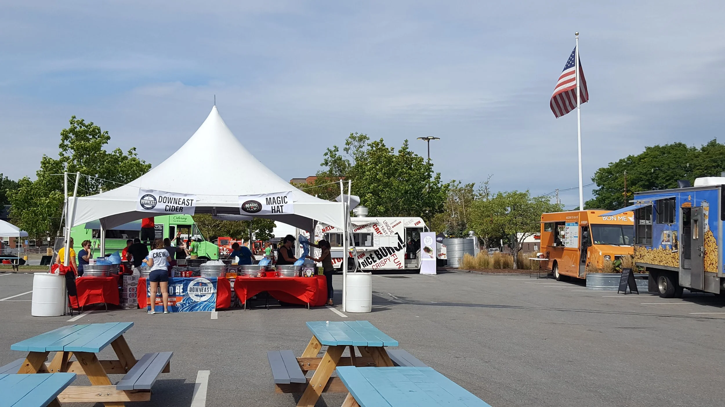 Food trucks and vendor tents in a parking lot, with people working at a food booth labeled 'Downeast Cider' and 'Magic Hat'. There are picnic tables in the foreground and a large American flag flying in the background.