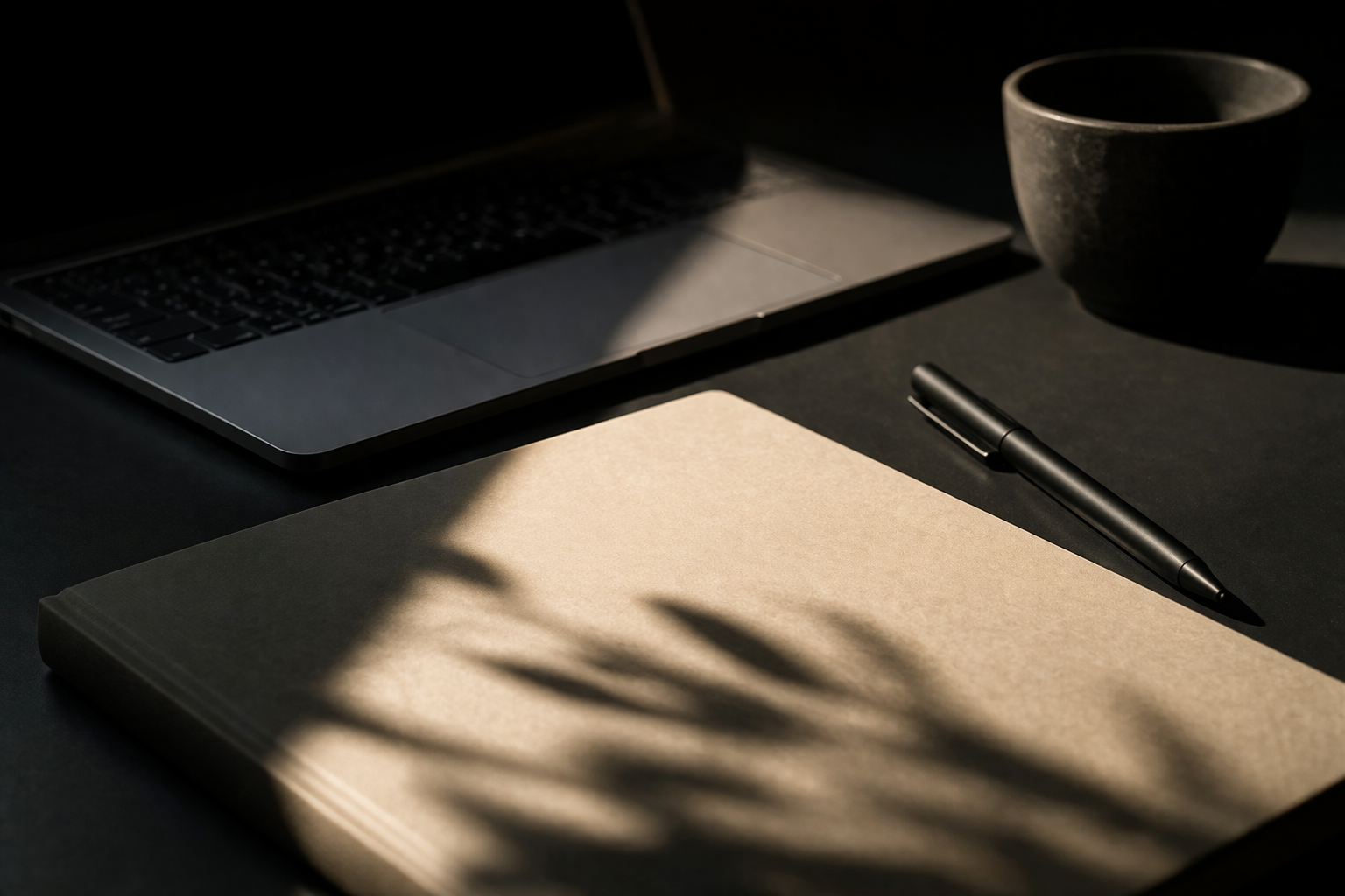 A workspace with a laptop, a closed notebook, a black pen, a ceramic cup, and a shadow of a plant on the table.
