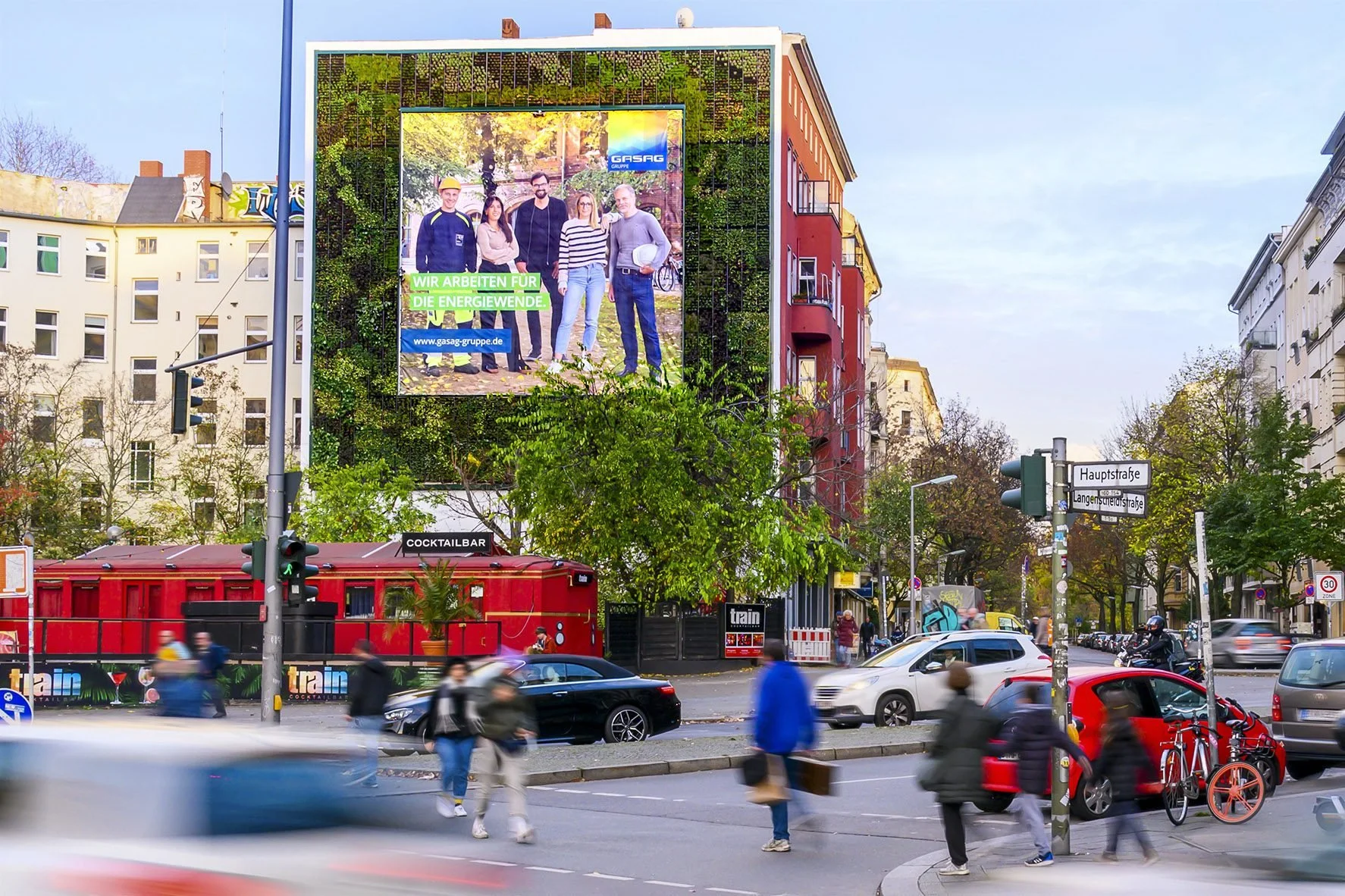 A busy city street corner with pedestrians crossing, cars, and a large digital billboard on a red building advertising a gas company, with trees and other buildings in the background.