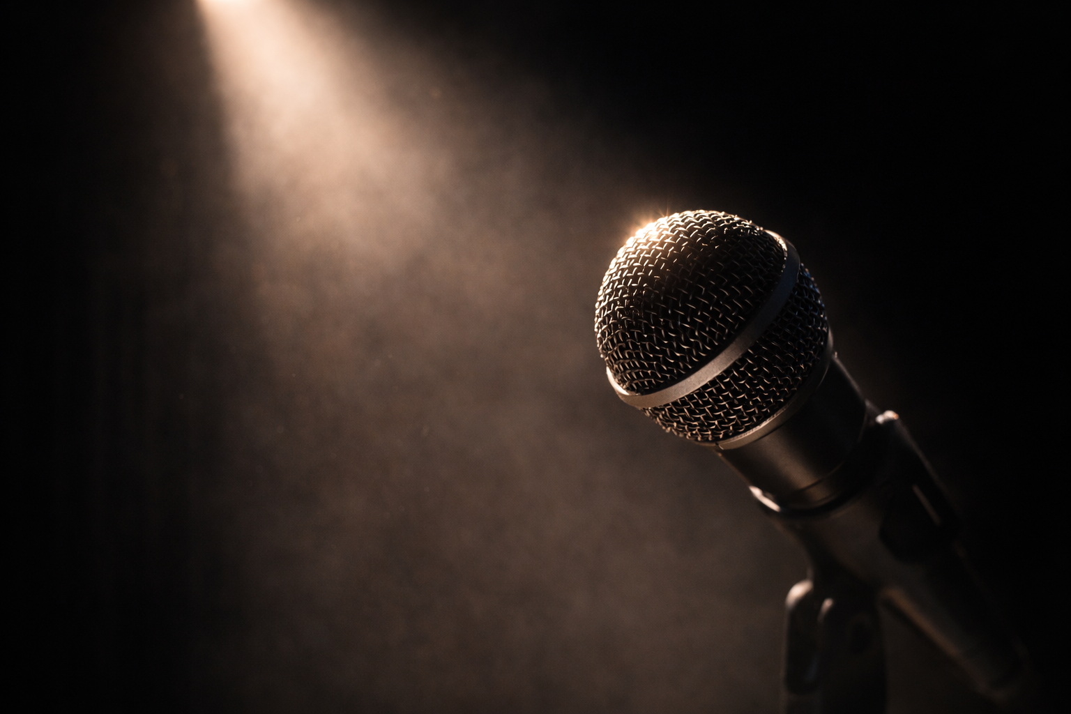 A close-up of a black microphone on a stand, lit by a single spotlight against a dark background.