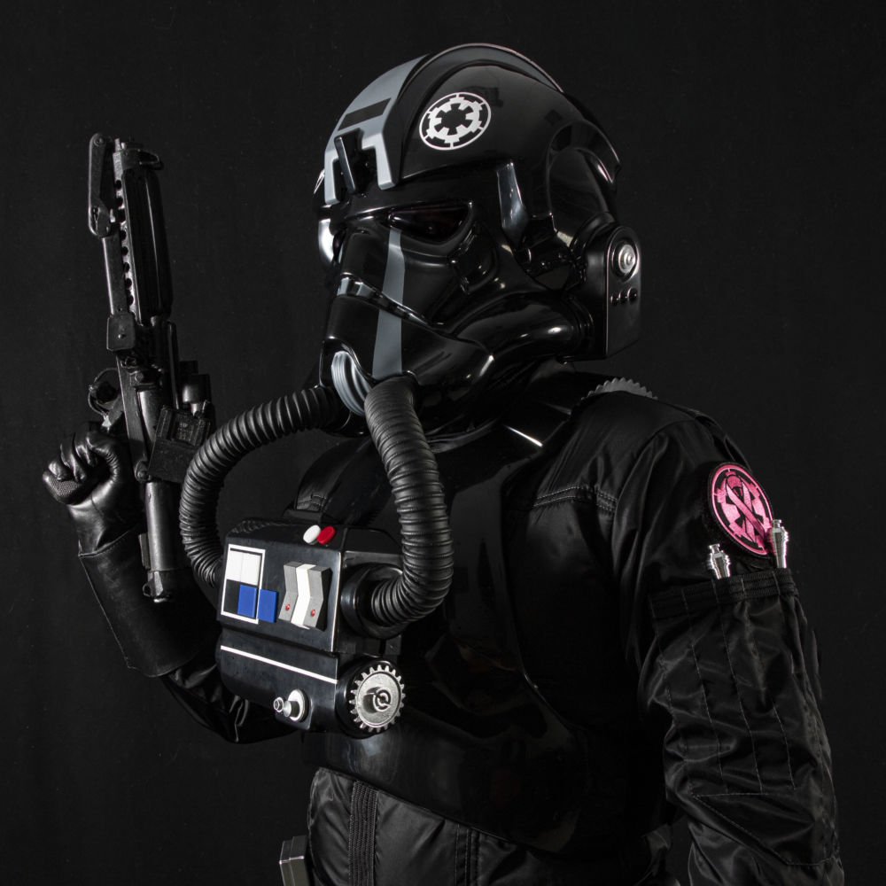 A person dressed as a Star Wars TIE fighter pilot in black uniform and helmet, holding a blaster against a plain black background.