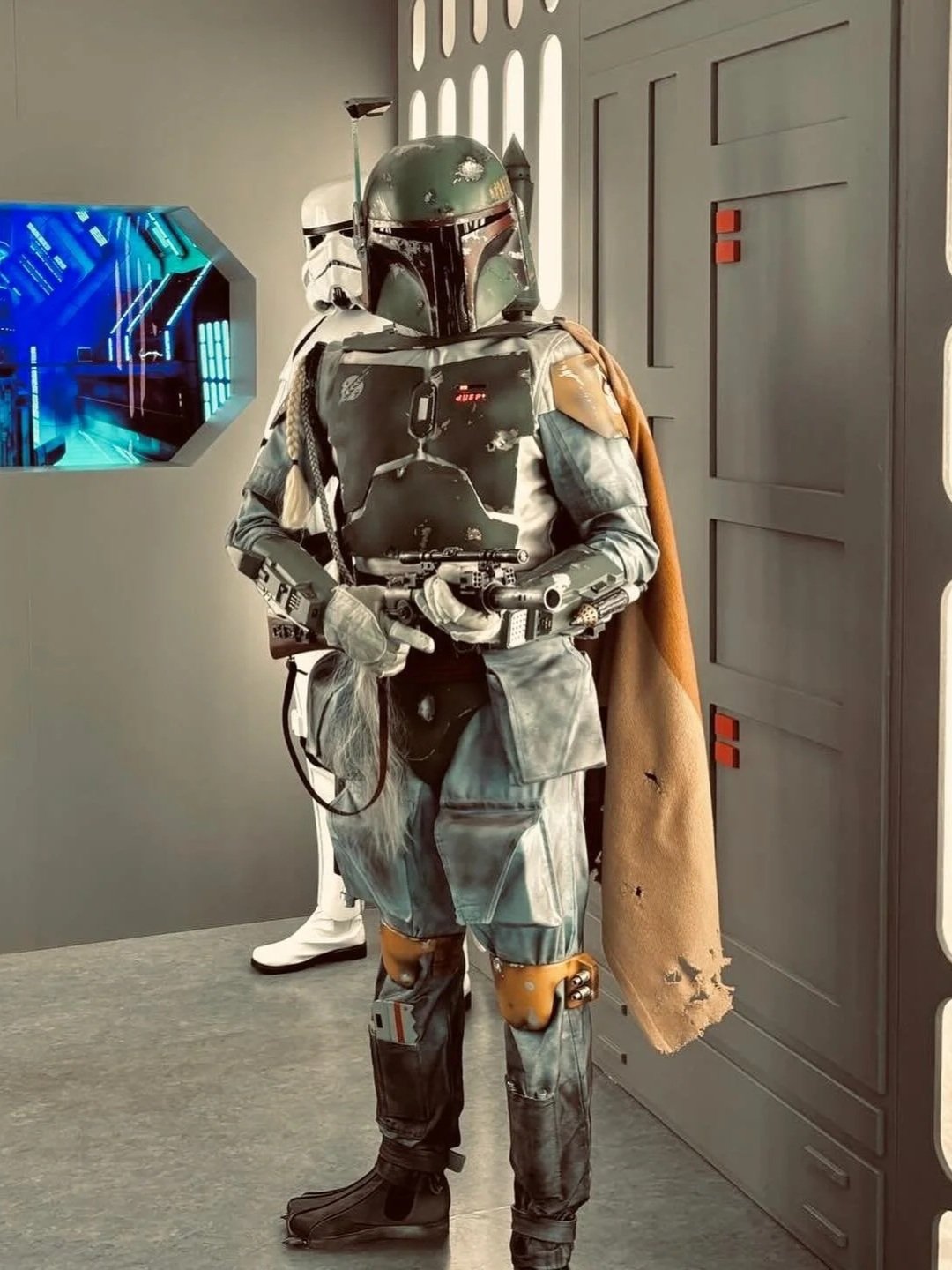 Star Wars Boba Fett collectible figure in a museum or display setting, dressed in armor with a helmet, holding a blaster, with a cape and detailed costume.