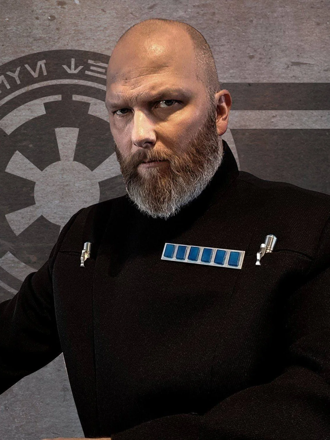 A man dressed as a Star Wars Imperial officer with a stern expression, standing in front of a gray wall with the Galactic Empire symbol.