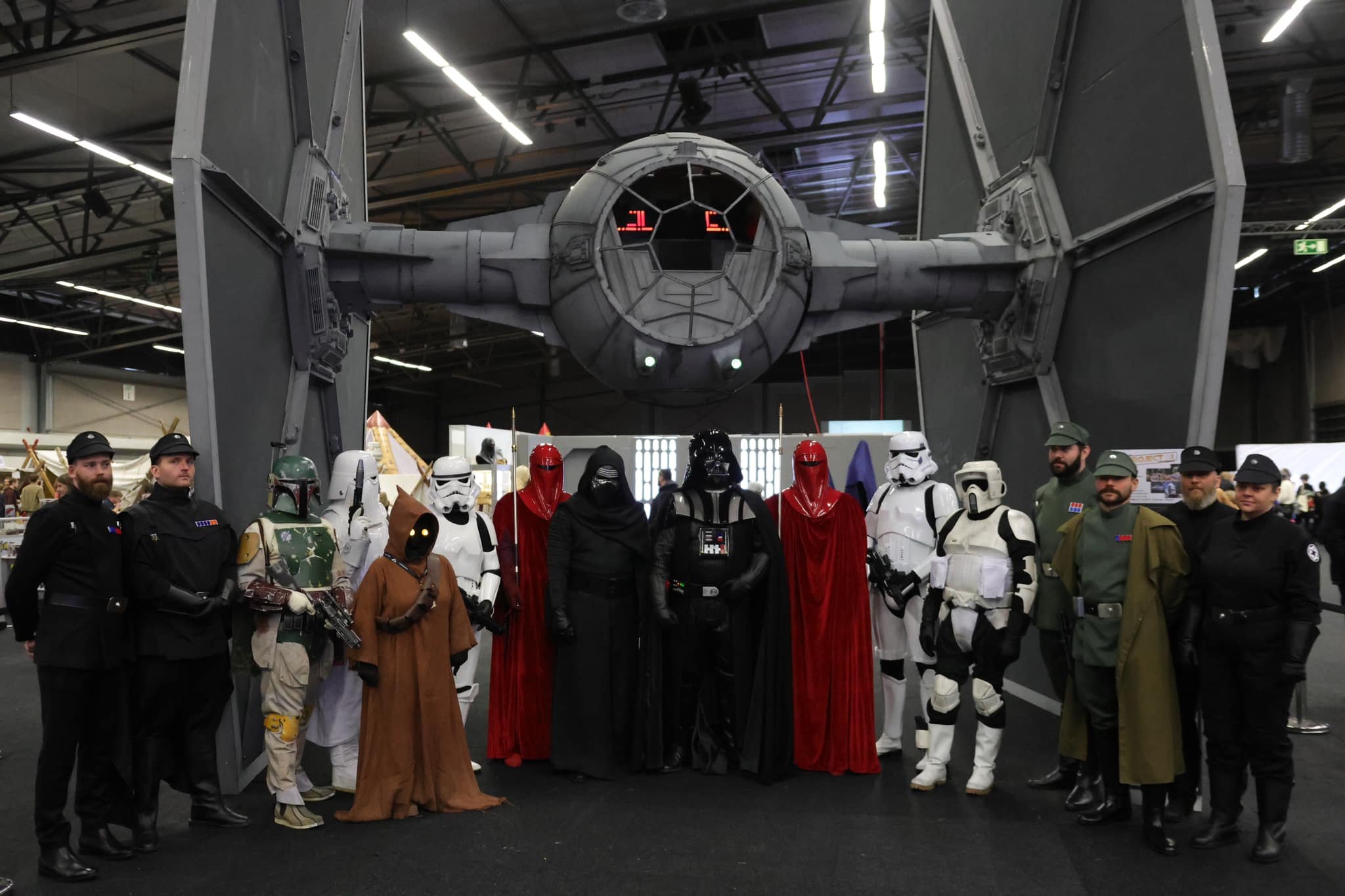Group of people dressed as Star Wars characters, including stormtroopers, Darth Vader, and others, standing in front of a replica of a Tie Fighter.