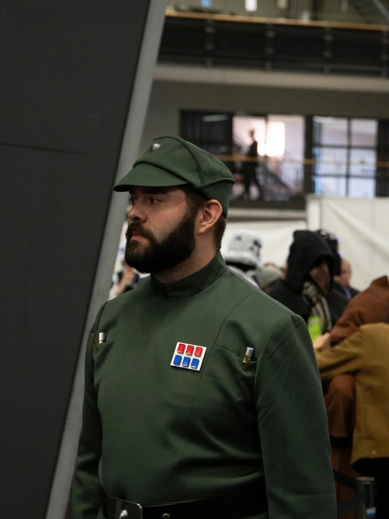 A man dressed as a Star Wars officer with a green uniform, cap, and medals, standing in a crowded indoor setting with people in the background.