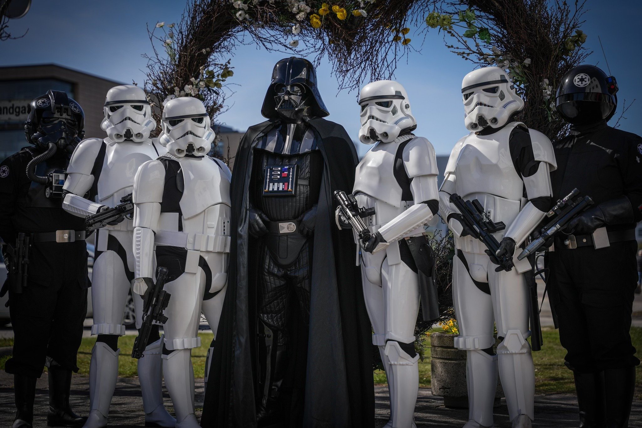 Group of people dressed in Star Wars costumes, including Darth Vader, stormtroopers, and officers, posing outdoors in front of a wreath and flowers.