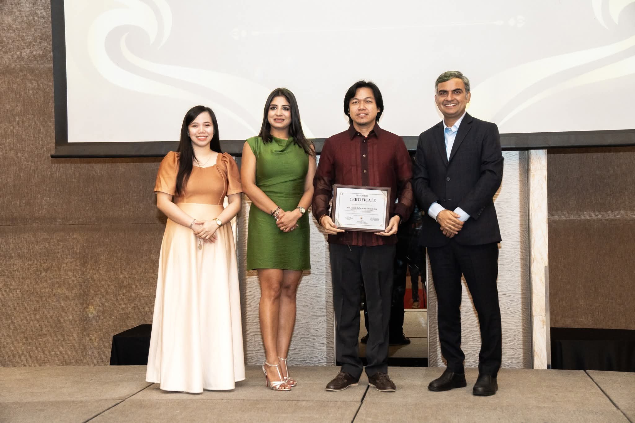 Four people stand on a stage, with one person holding a certificate, in front of a large screen at an award ceremony.