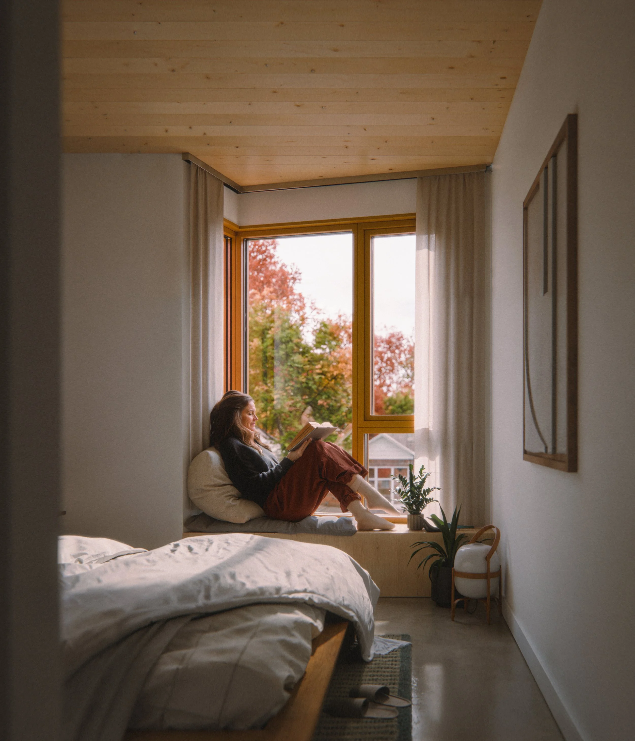 A woman sitting on a window seat in a bedroom, reading a book. The room has a wooden mass timber ceiling, beige curtains, and a window with an autumn landscape outside. There are potted plants on a stand beside her bed.