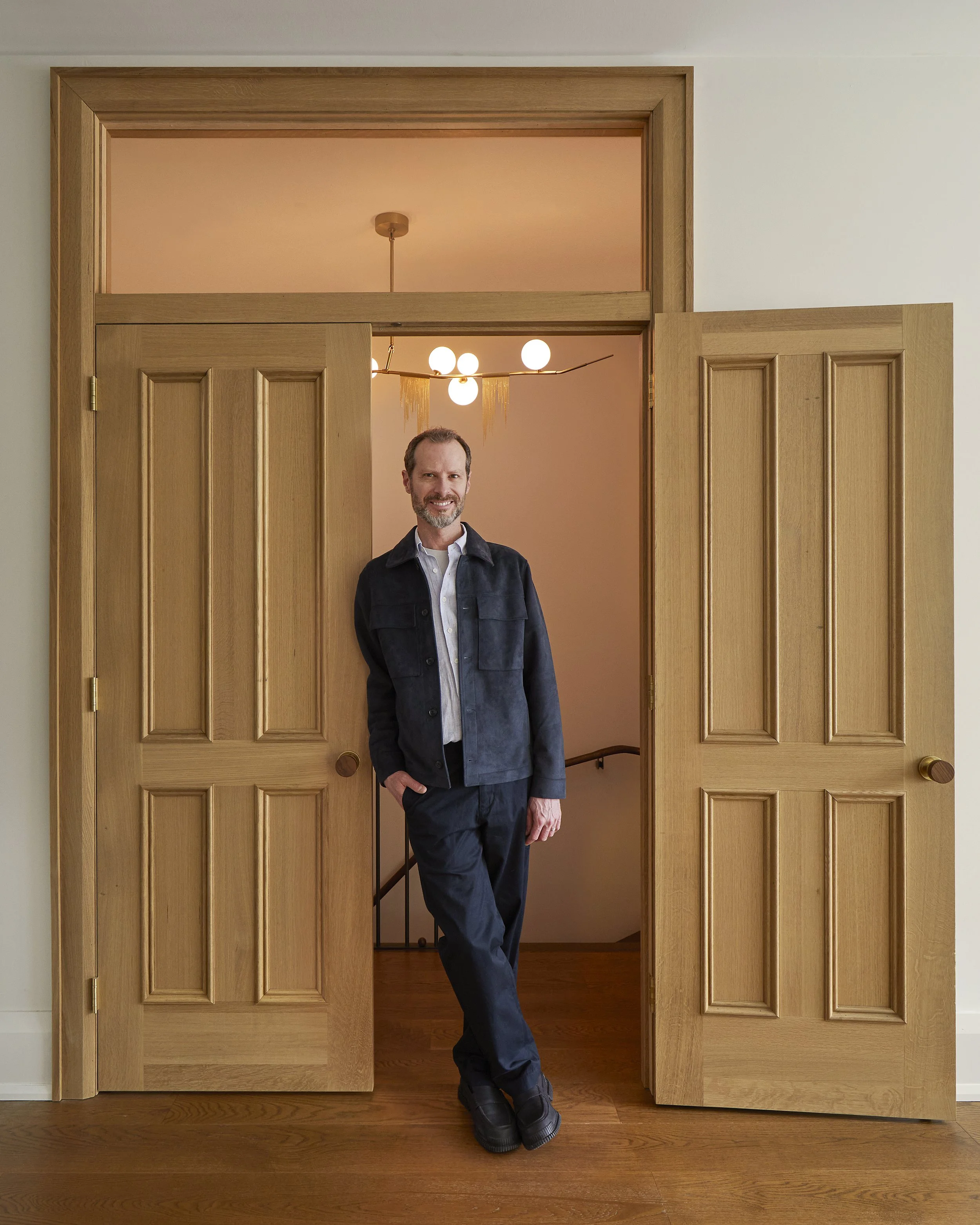 A man with a beard standing in an open doorway, wearing dark clothing, smiling, with a modern chandelier hanging inside the room behind him, and a staircase visible in the background.