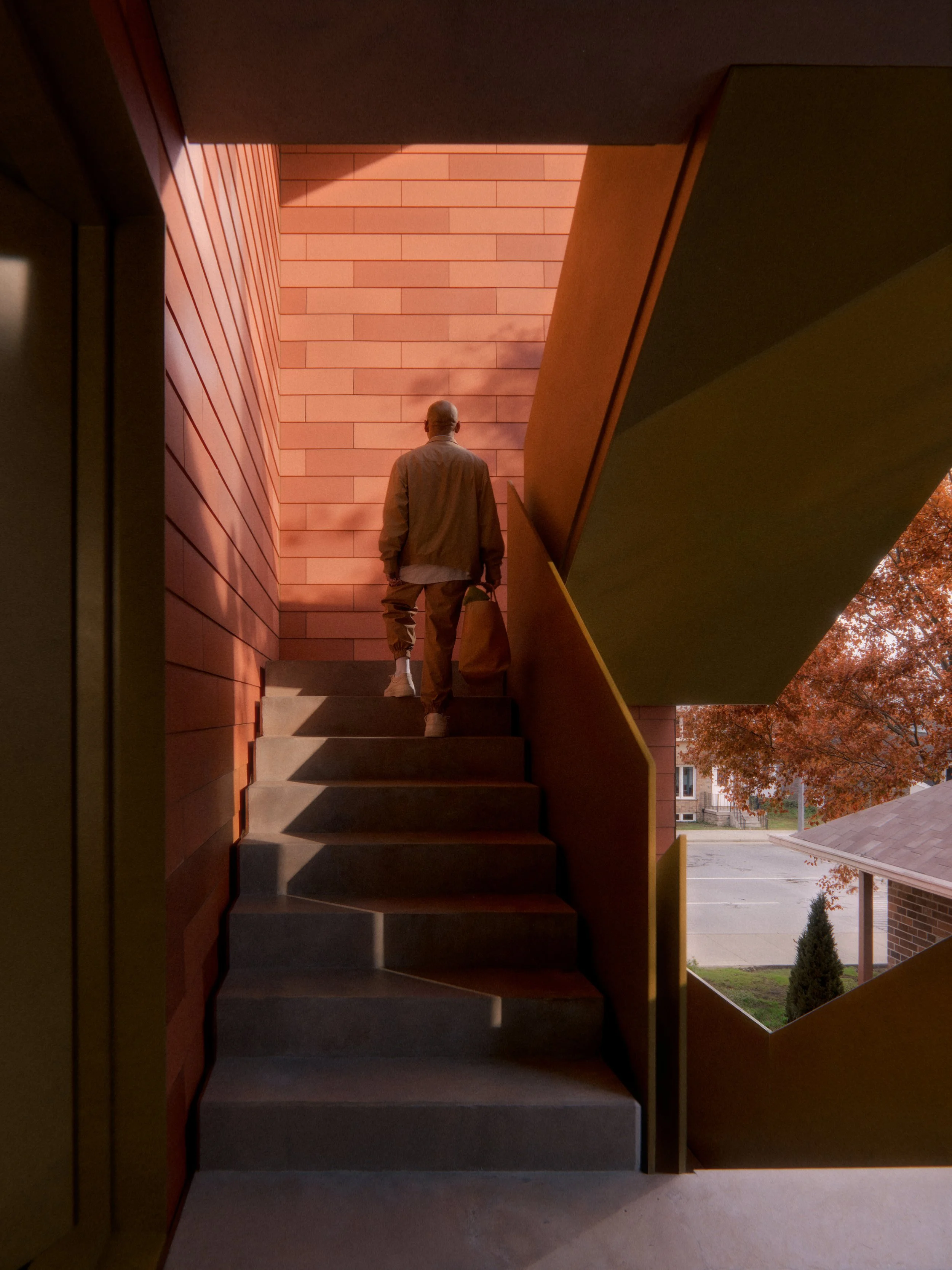 A man with a bag walking up an outdoor single egress staircase at sunset, with modern architectural elements and autumn trees in the background.