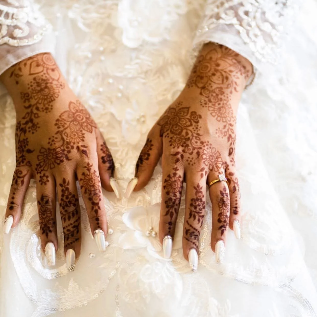Close-up of hands with intricate henna tattoos on fingers and back of hands, resting on a white lace fabric, with white nail polish and a gold ring.