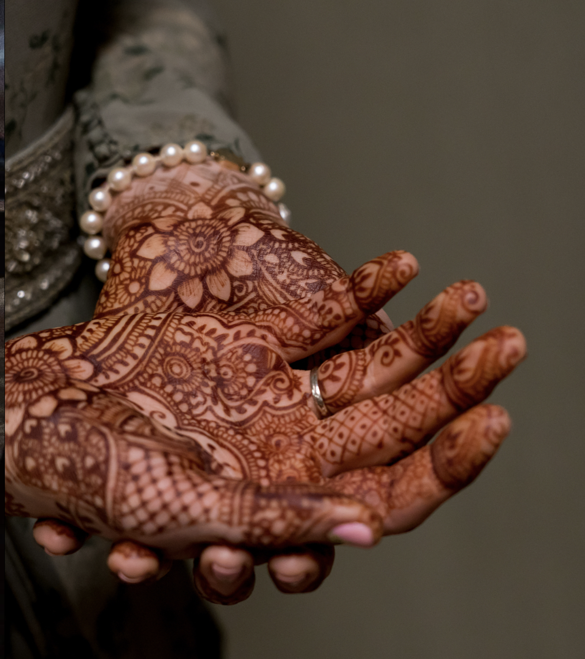 Close-up of hands decorated with intricate henna designs, wearing a pearl bracelet and wedding ring, with a blurred background.