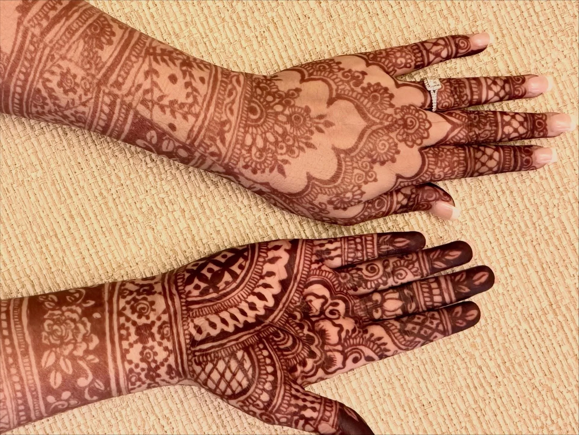 Two hands decorated with intricate henna designs, placed on a woven mat.