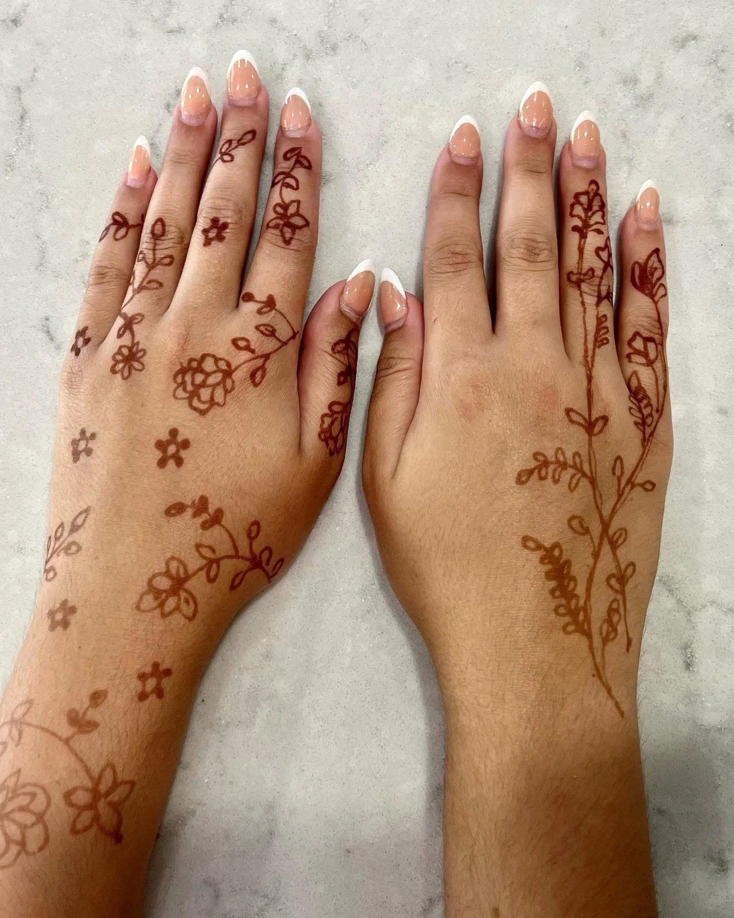Two hands with henna designs placed on a light surface, showing floral and vine patterns on the palms and fingers, with neatly filed nails painted with French manicure.
