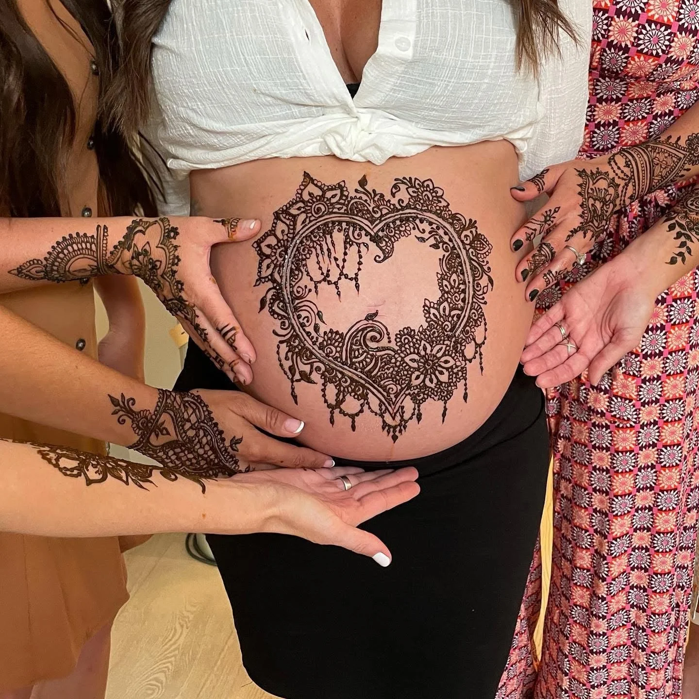 A pregnant woman with a decorative tattoo on her belly, surrounded by four people with various tattoos on their arms who are gently touching her belly.
